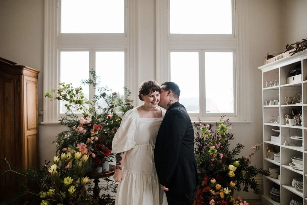 A couple stands close together in front of large windows, smiling and touching foreheads, with floral arrangements behind them in a bright room.