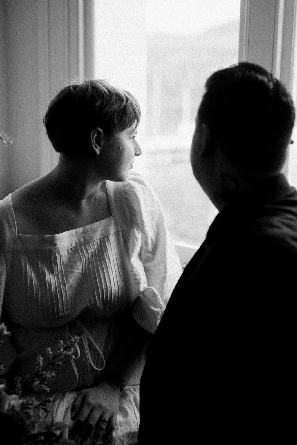 A black-and-white photo of a woman and a man sitting close together by a window, facing each other, with the woman looking out the window and smiling softly.
