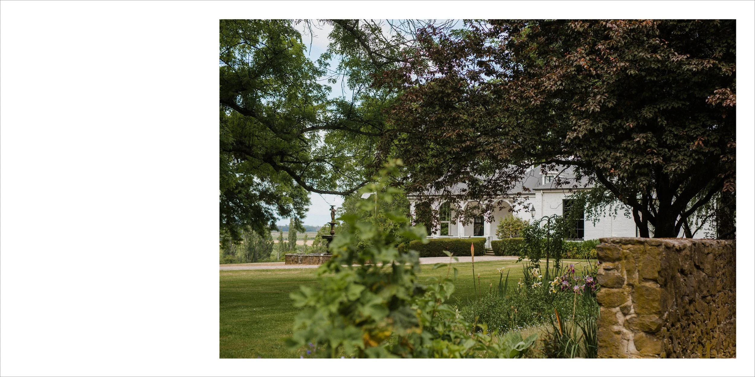 A white house with a landscaped garden and large trees, including a stone wall in the foreground, a fountain, and a grassy lawn with flowers.