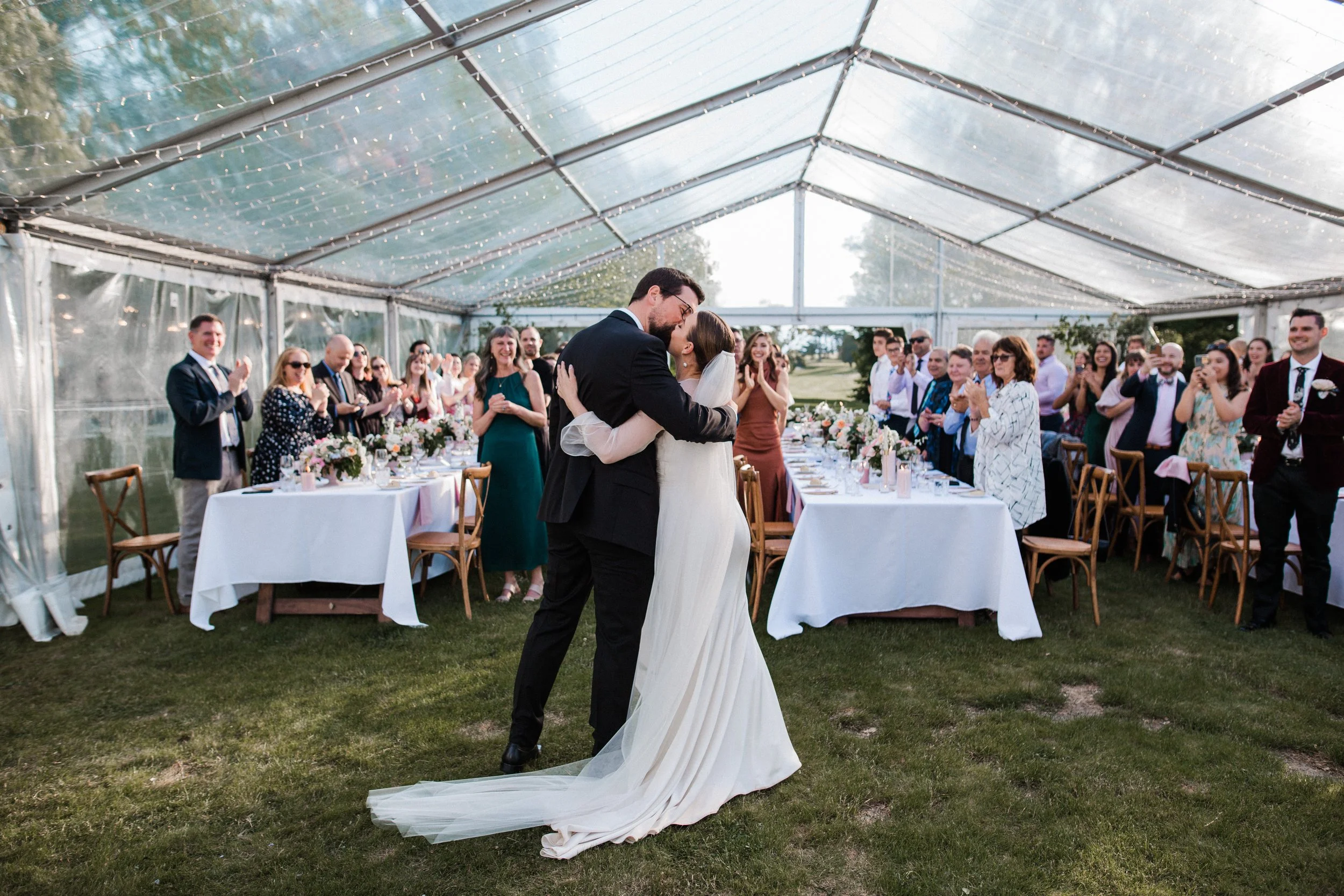 A newlywed couple sharing a dance under a large transparent tent, surrounded by seated guests clapping and smiling at the wedding reception outdoors.