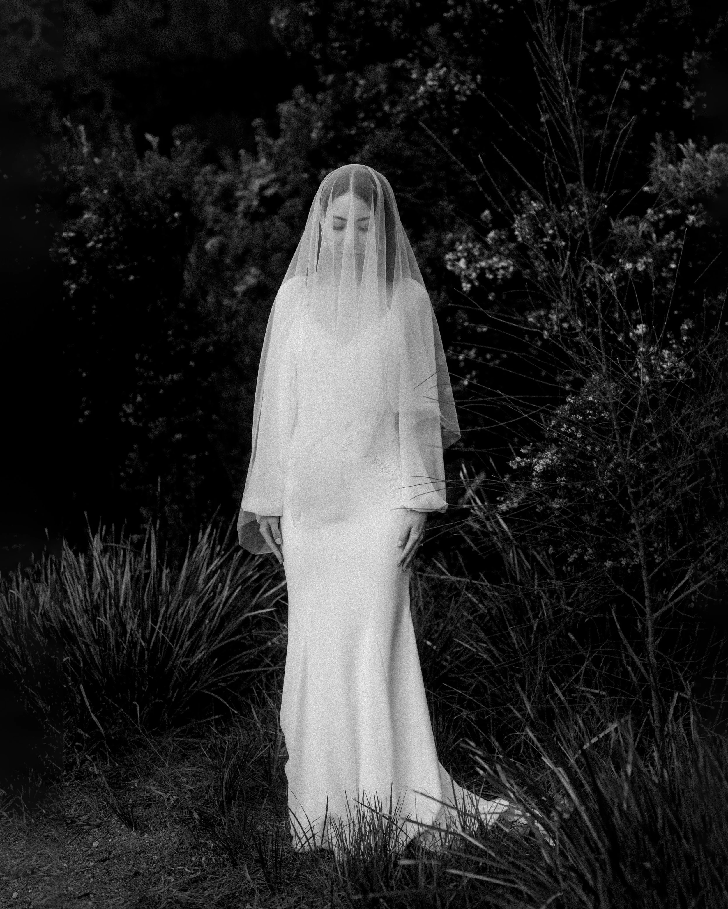 A woman dressed in a long white gown with a veil covering her face, standing outdoors among grass and bushes, captured in black and white.
