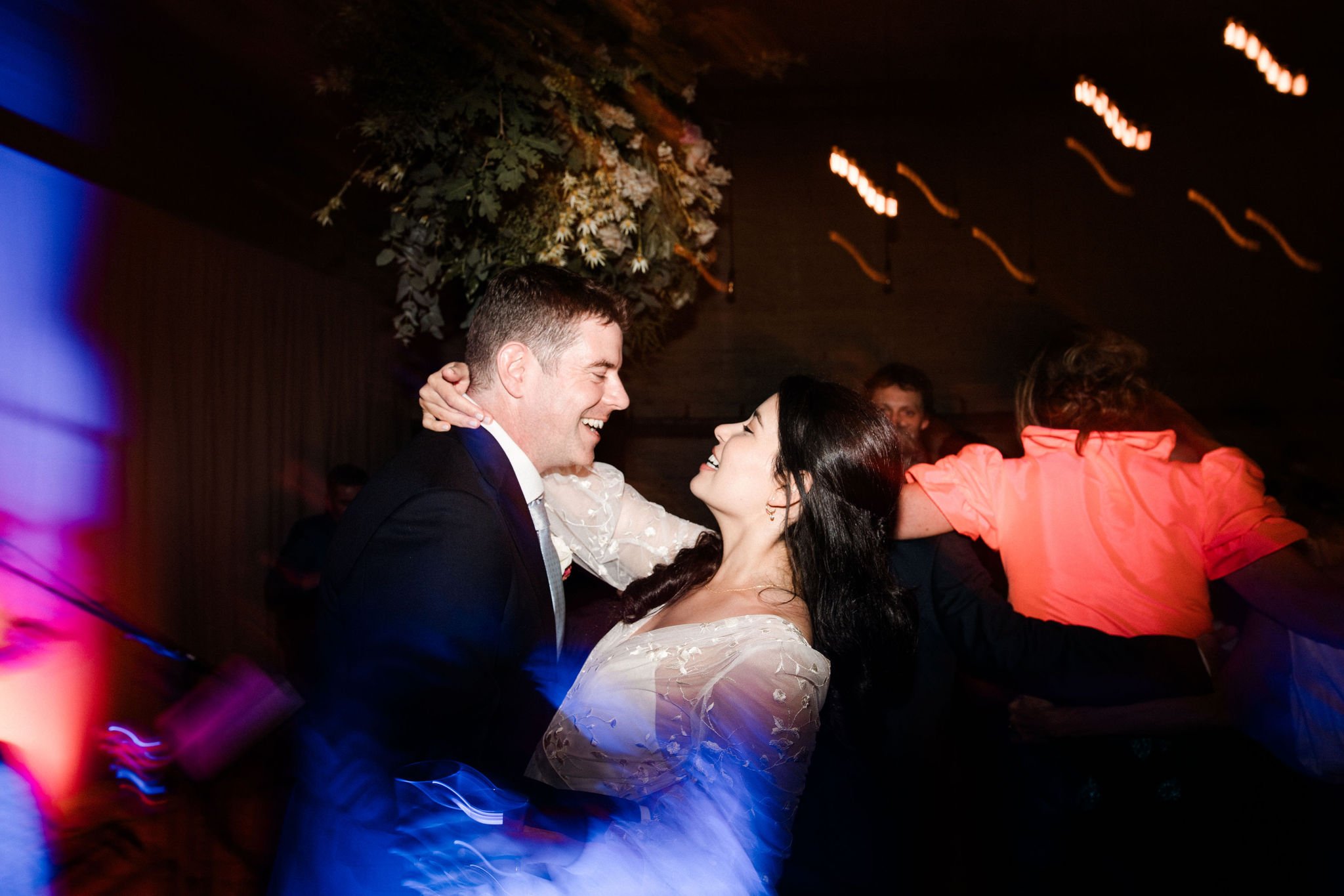 A couple dances closely and happily at a celebration, with a man in a suit and a woman in a white dress, while other people dance in the background.