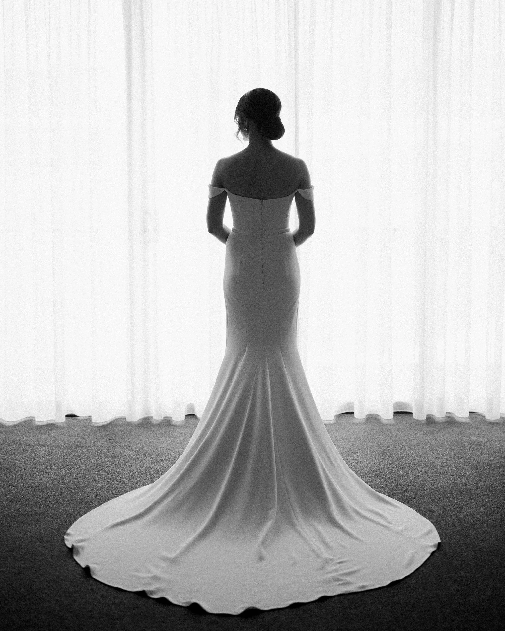 Black and white photo of a woman in a wedding dress standing in front of a sheer curtain. She is facing away, with her hair styled in an updo, and the dress has off-shoulder sleeves and a train.