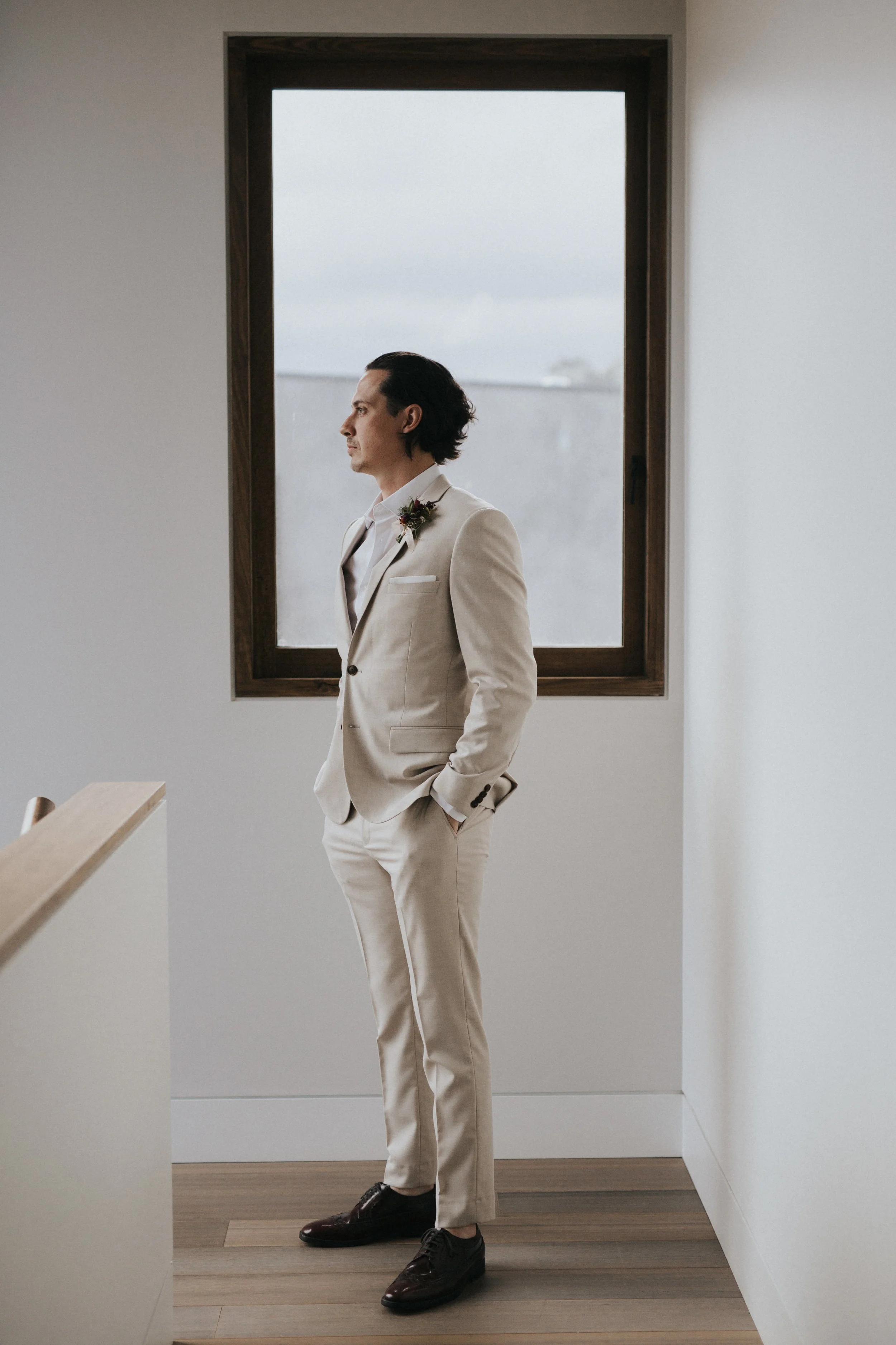 A man dressed in a light-colored suit with a boutonniere on his lapel, standing indoors near a large window with his hands in his pockets, looking out contemplatively.