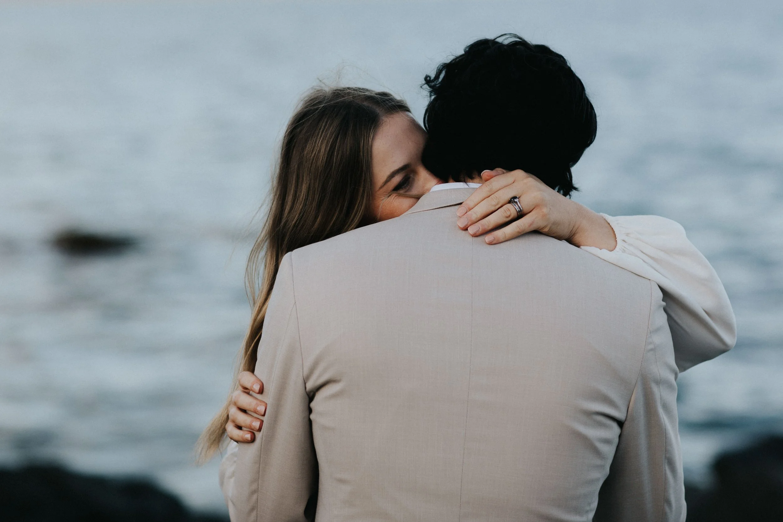 A woman with long light brown hair hugging a man with dark hair near a body of water at Piermont, Tasmania..