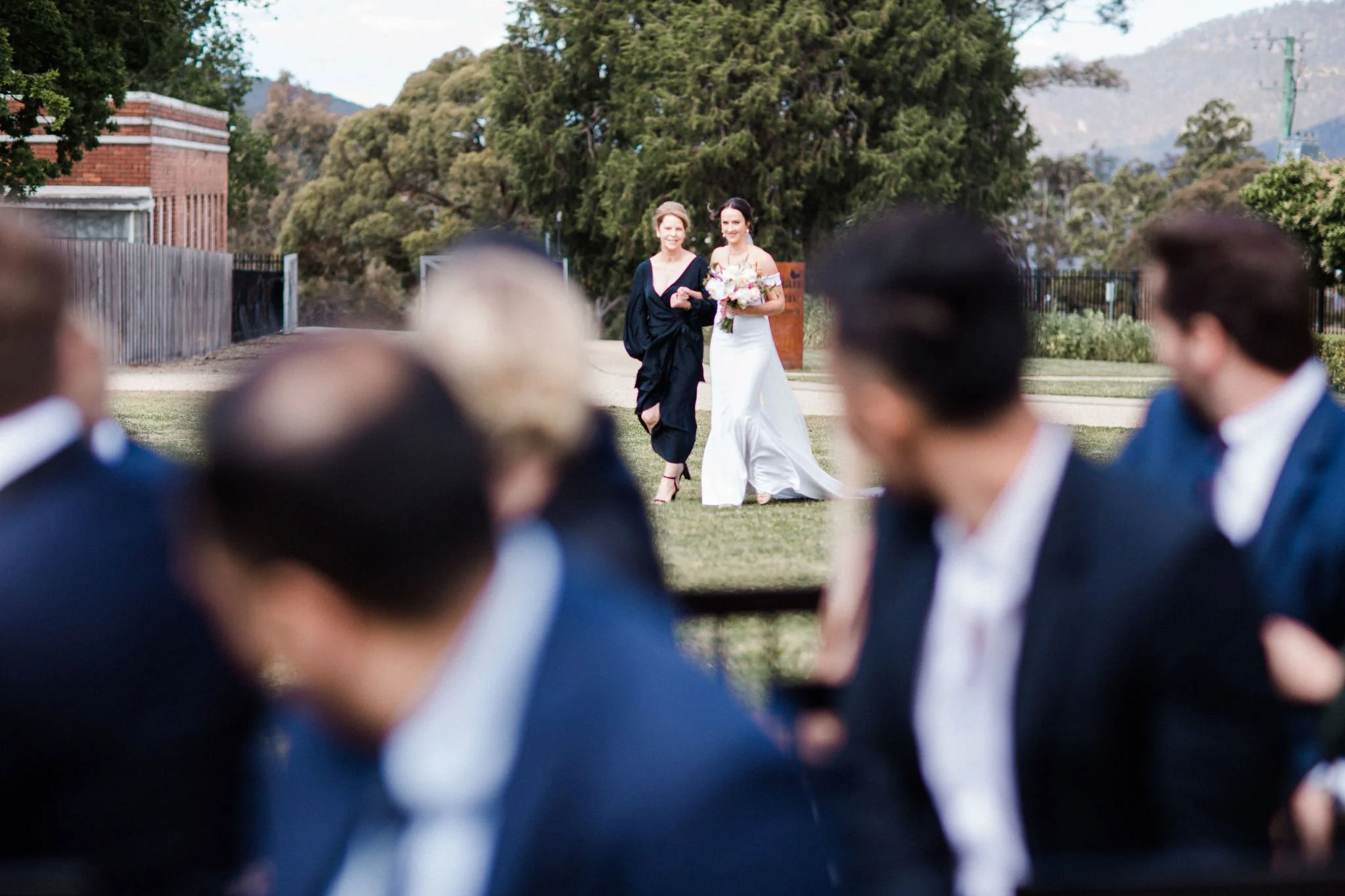 Bridesmaid walking with bride down aisle at outdoor wedding, blurred guests in foreground, trees and a building in background.