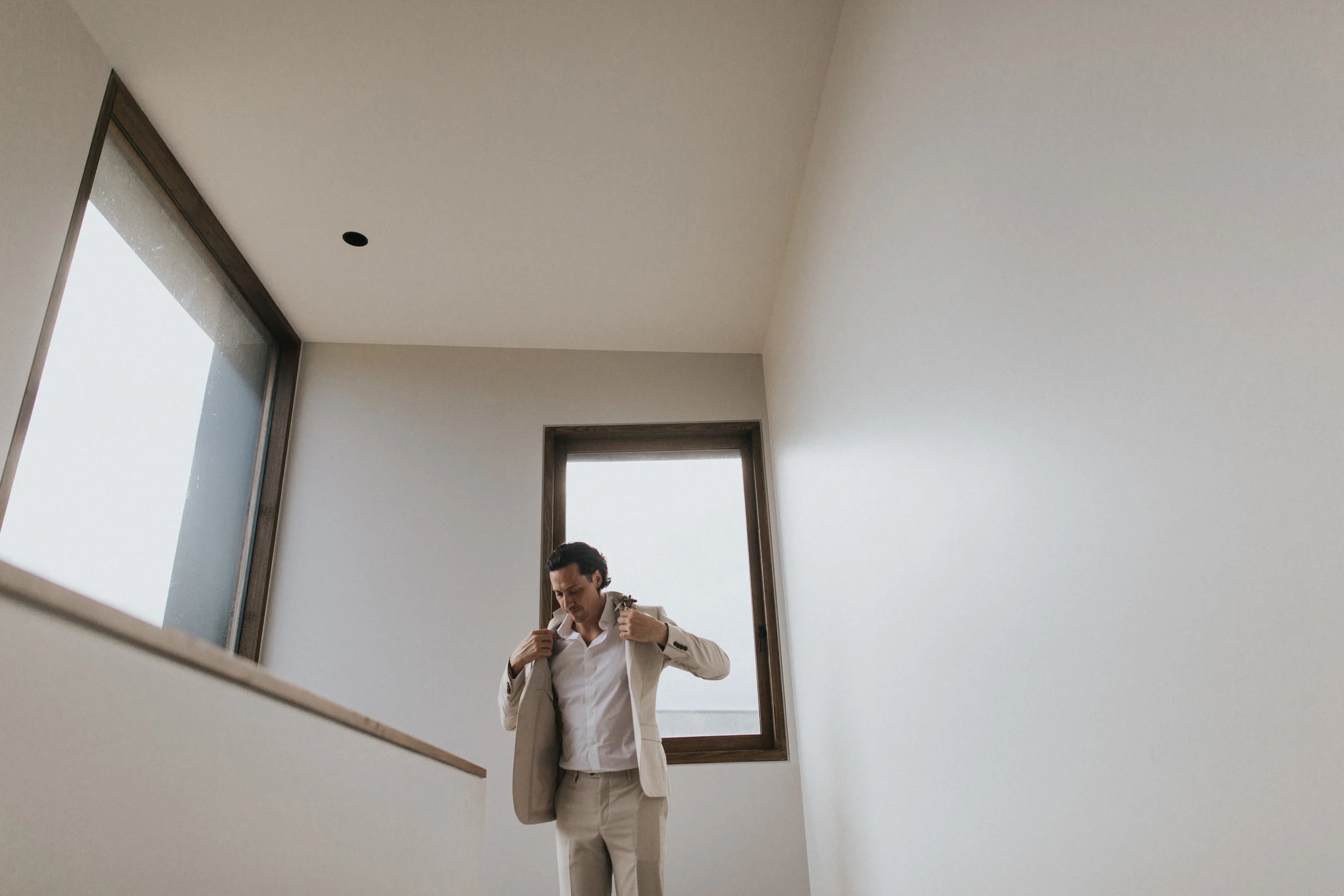 Man in a beige suit putting on a jacket in a minimalist room with white walls and large windows.