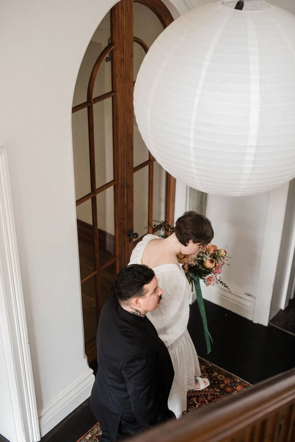A couple standing in an entryway, the woman holding a bouquet of flowers, with a large paper lantern hanging overhead.