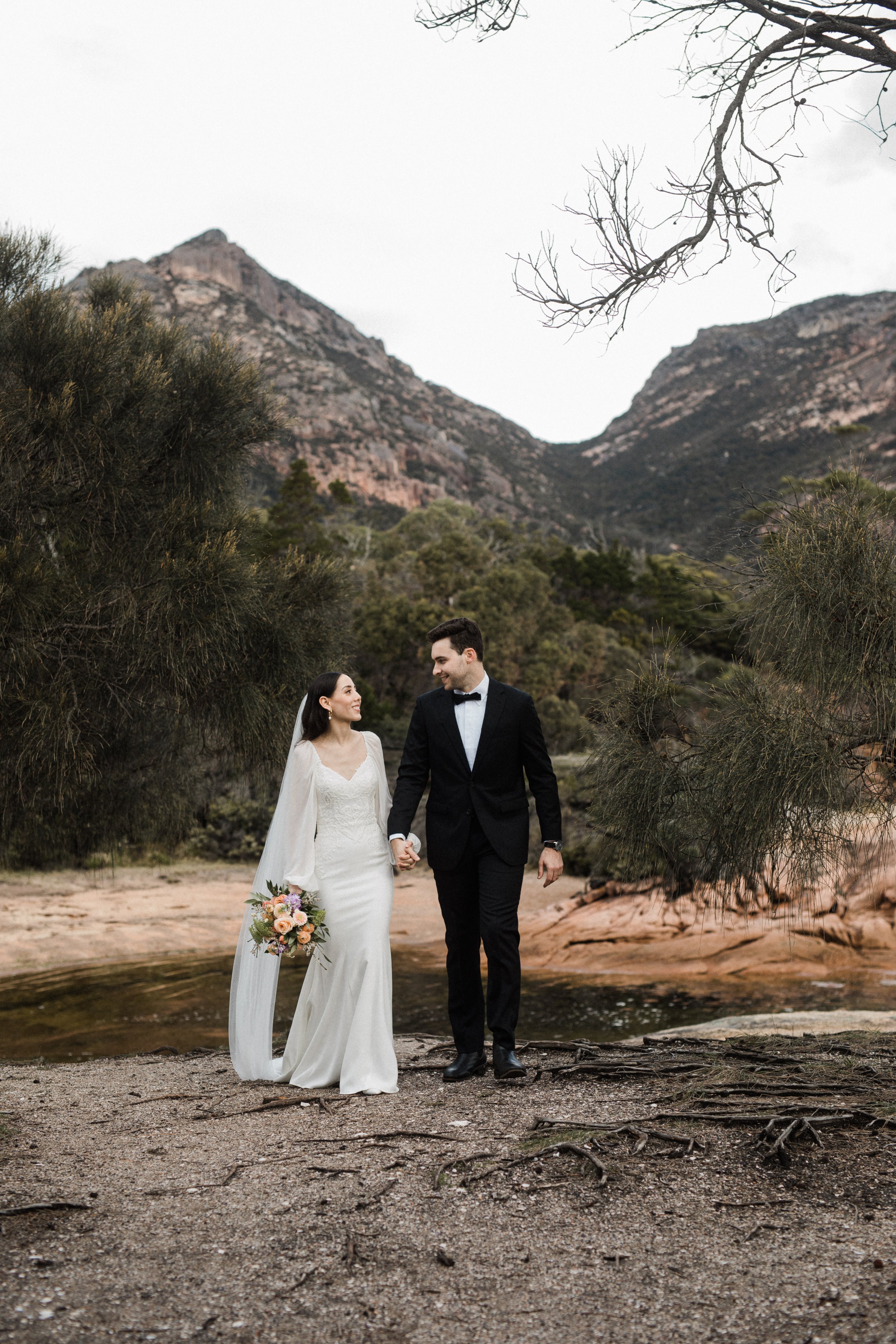 A bride and groom holding hands and walking outdoors near a small stream with mountains and trees in the background on a cloudy day.