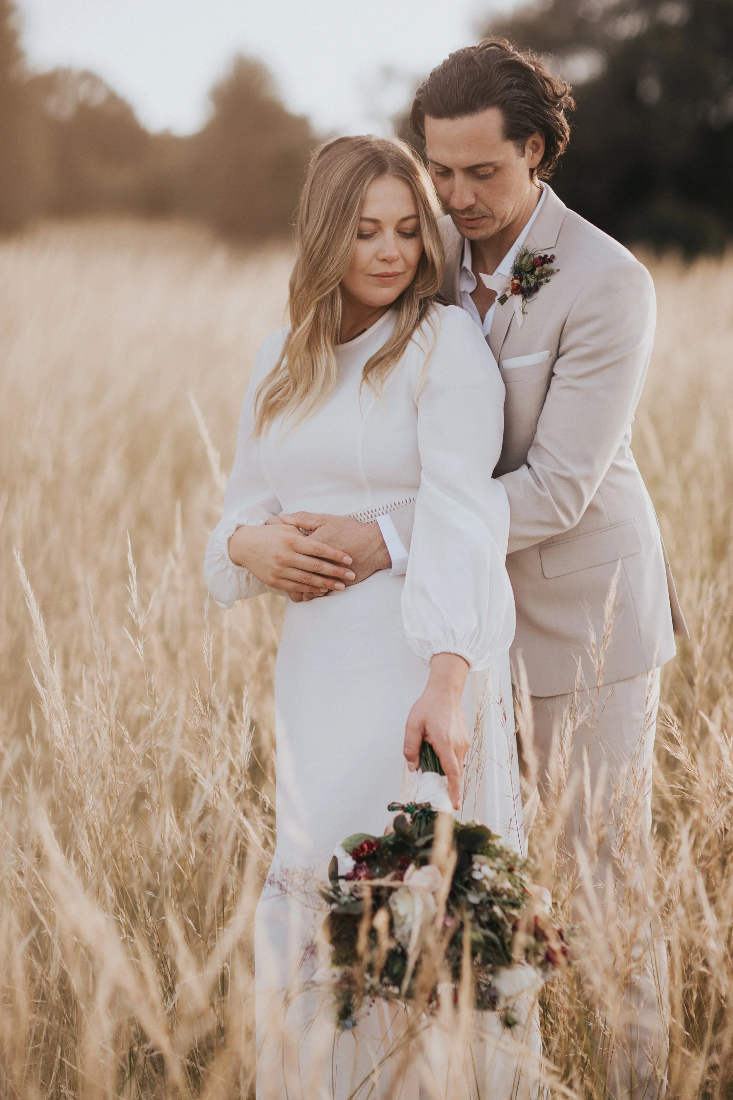 A man and woman holding each other in a field of tall, golden grass, with the woman holding a bouquet of flowers.