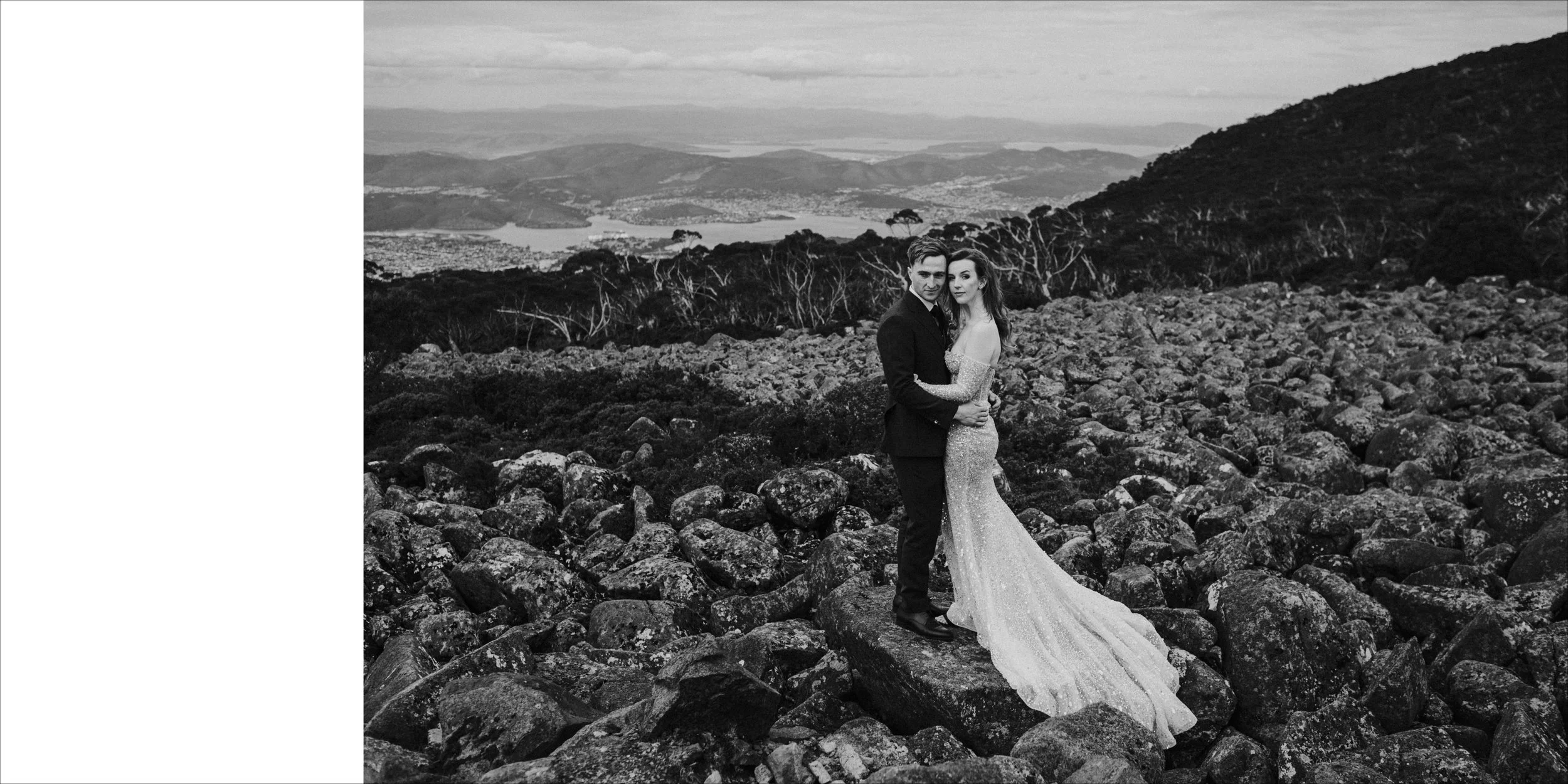 Black and white photo of a bride and groom standing on rocky terrain with a mountain and lake landscape in the background