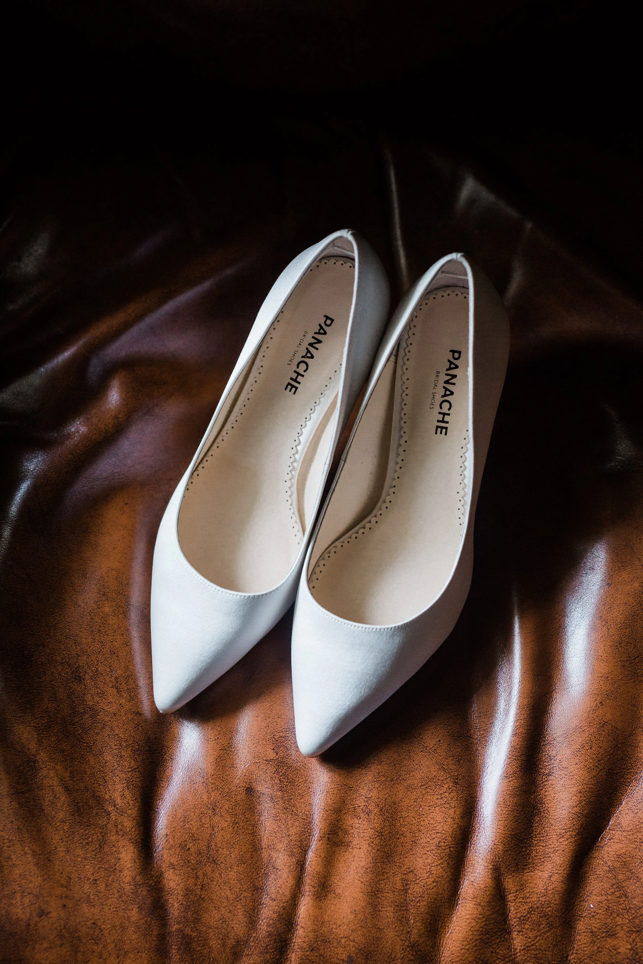 Pair of white pointed-toe high heels on brown leather surface.