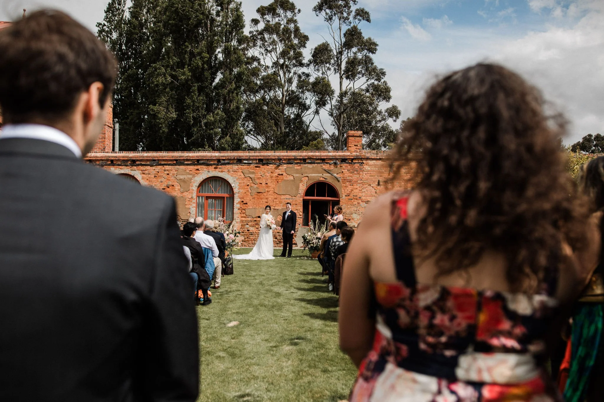 A wedding ceremony outdoors on green grass with a brick building and trees in the background, where a bride and groom stand together in front of seated guests.