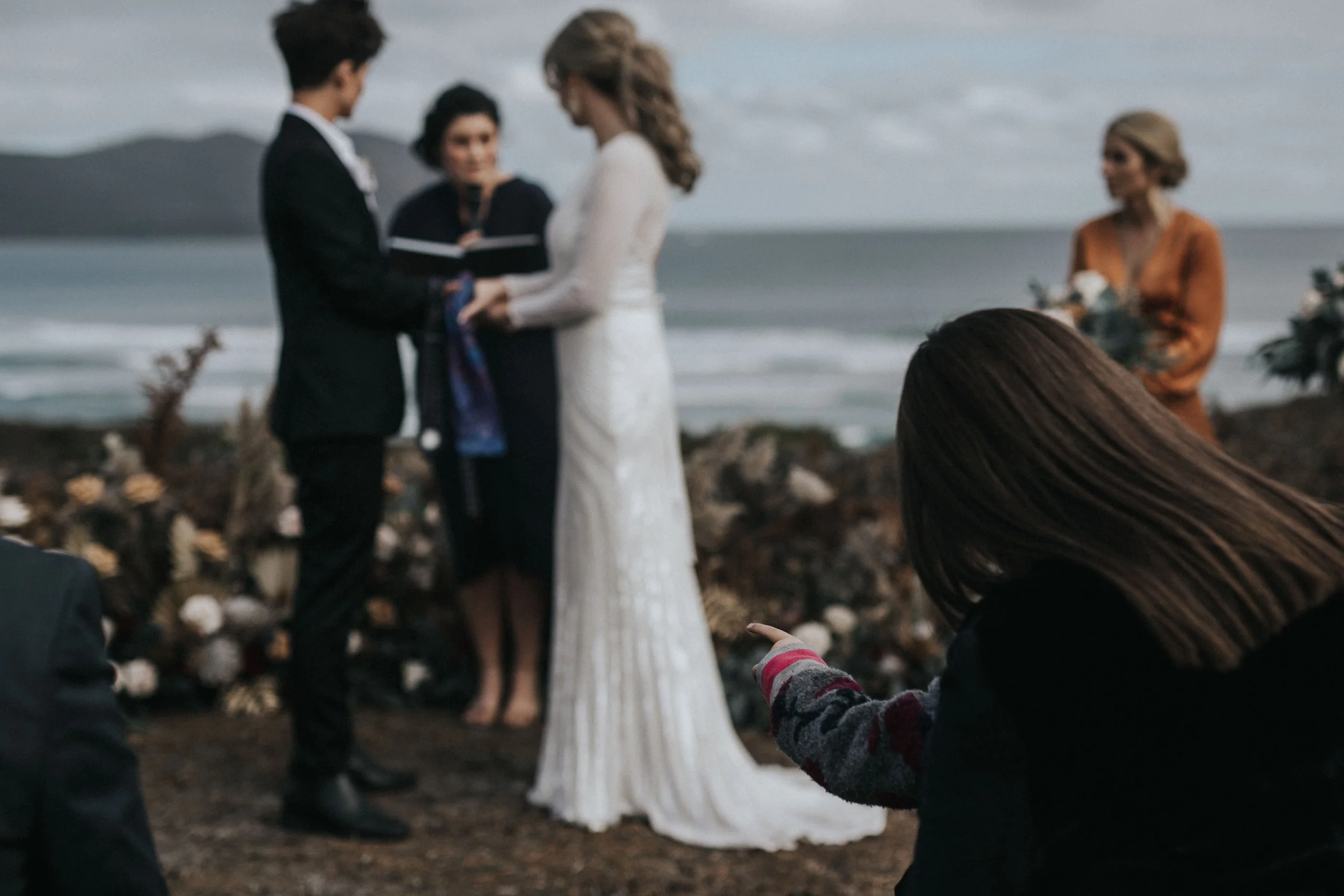 A beachside outdoor wedding ceremony with a bride and groom exchanging vows, an officiant, an additional woman holding flowers, and a girl pointing towards the couple in the foreground.