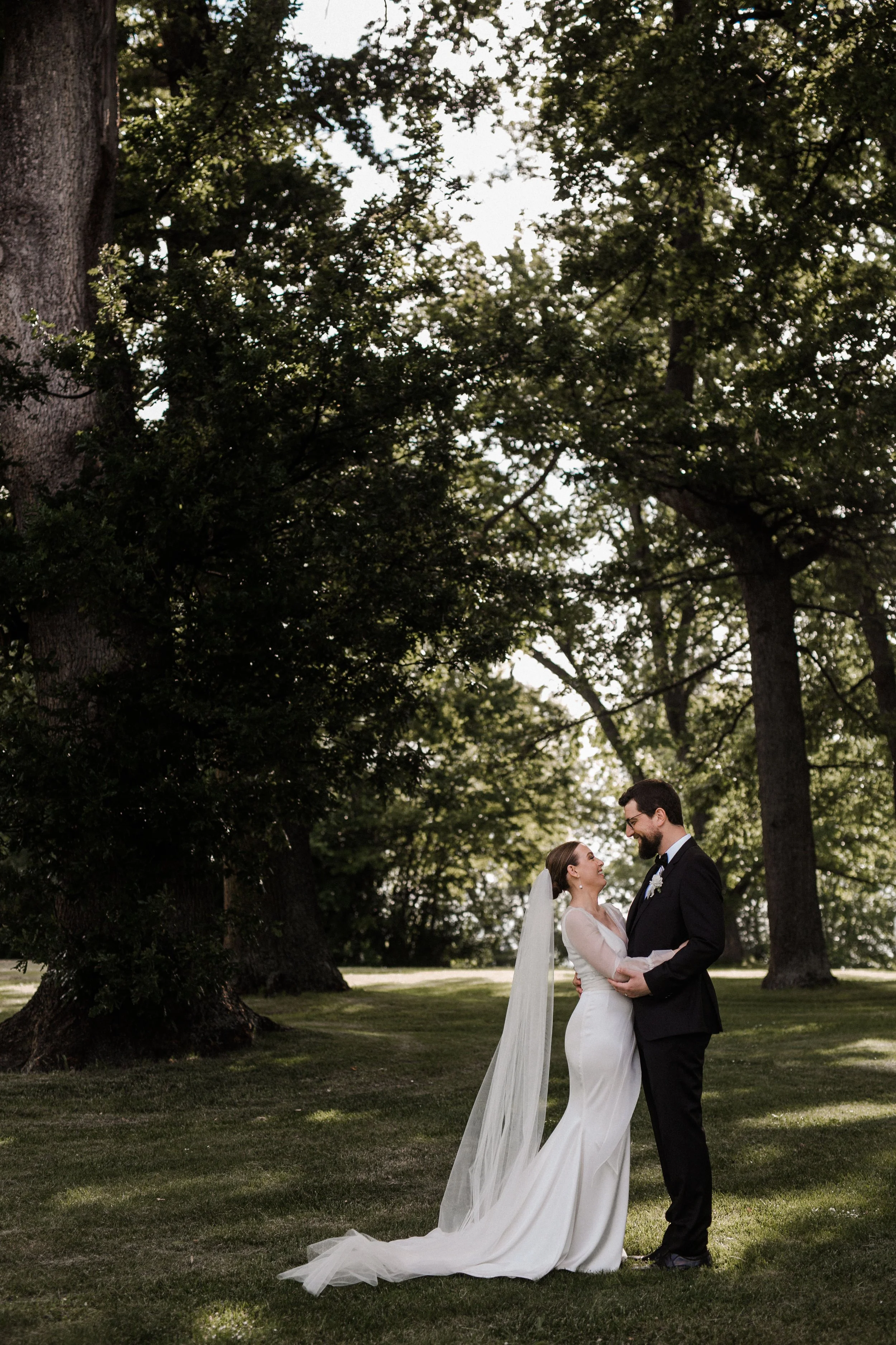 A bride and groom standing outdoors on grass, holding hands and looking at each other, surrounded by large trees with green leaves on a sunny day.
