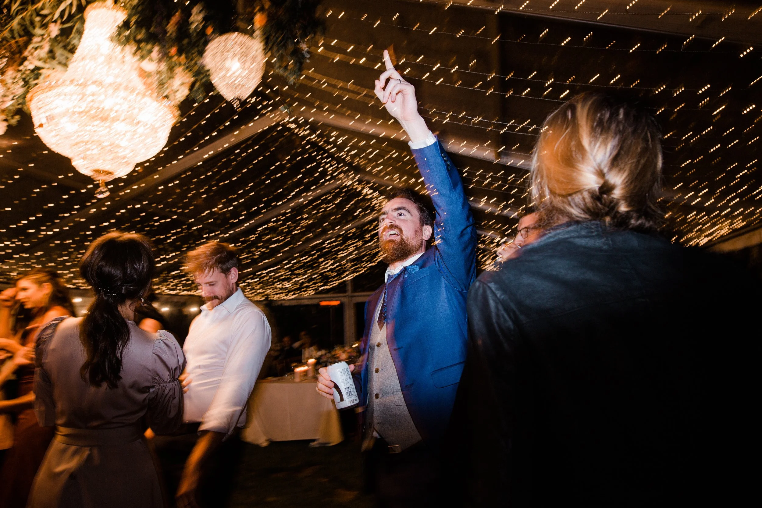 People dancing and socializing at a wedding reception under hanging string lights and chandeliers.