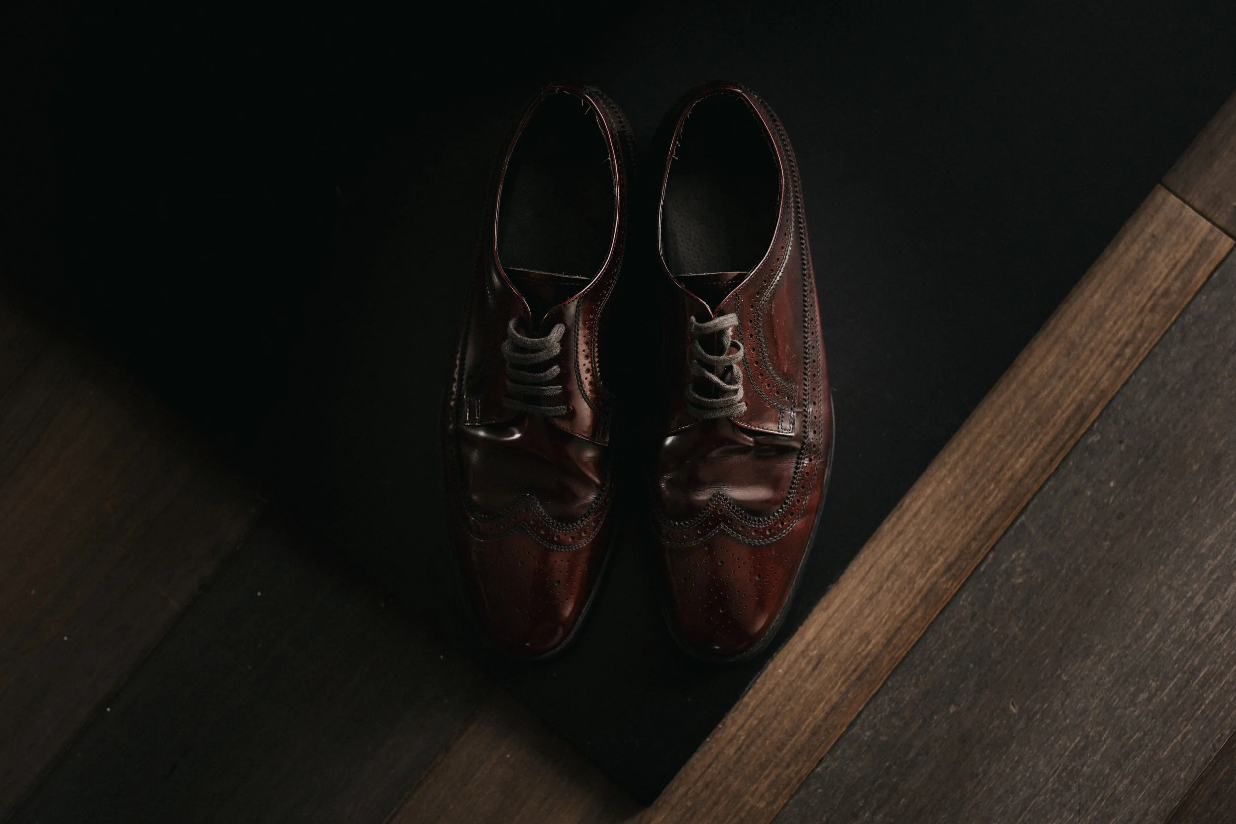 Pair of polished brown leather dress shoes with brogue details, placed on a dark surface against a dark background and wooden flooring.