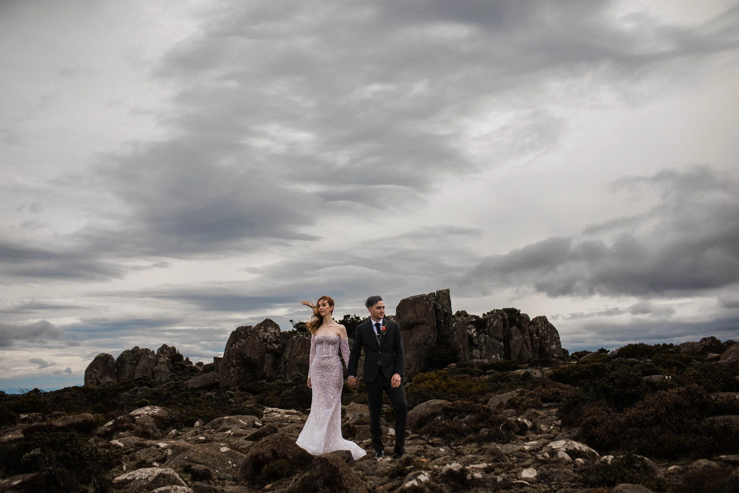 A bride and groom walking hand in hand on a rocky landscape with large rocks and bushes, under a cloudy sky.