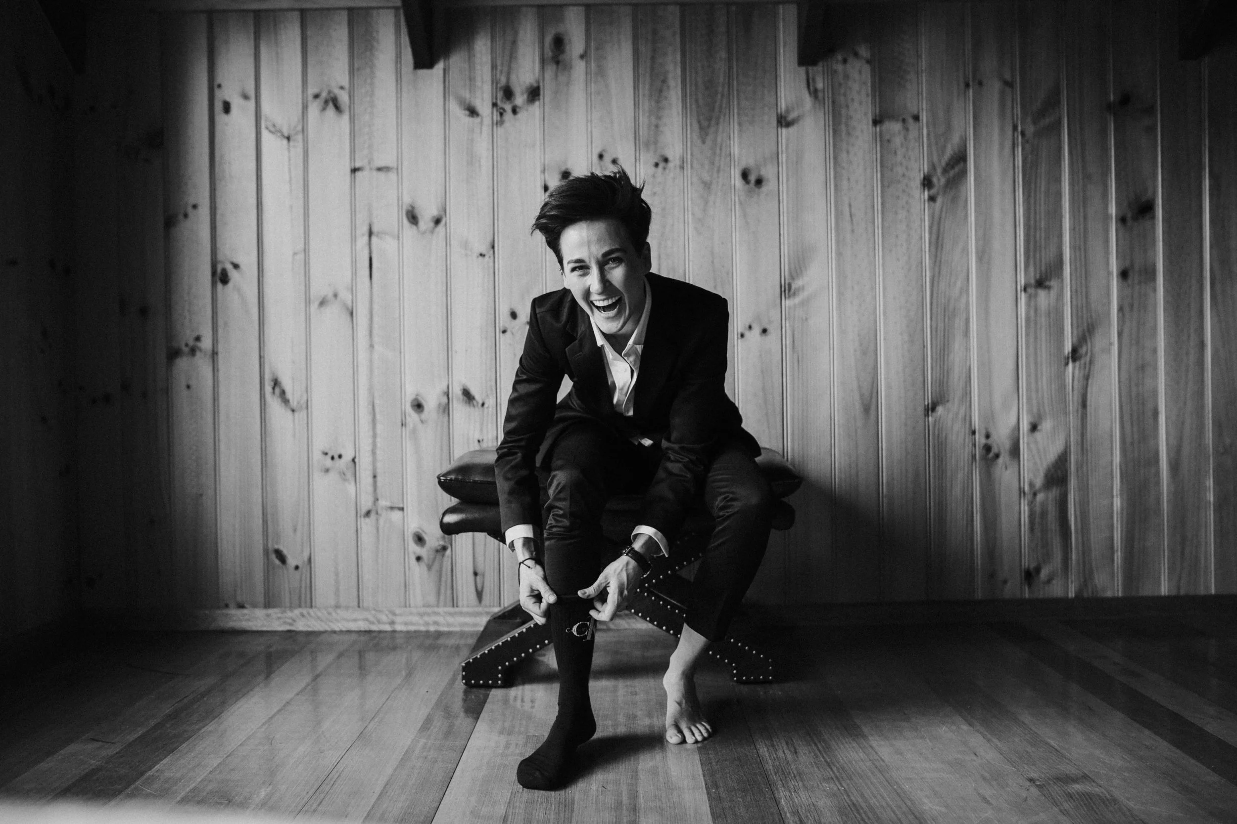 Black and white photo of a woman in a suit sitting on a bench, laughing, as she adjusts her sock and shoe, in a room with wooden walls and hardwood floor.