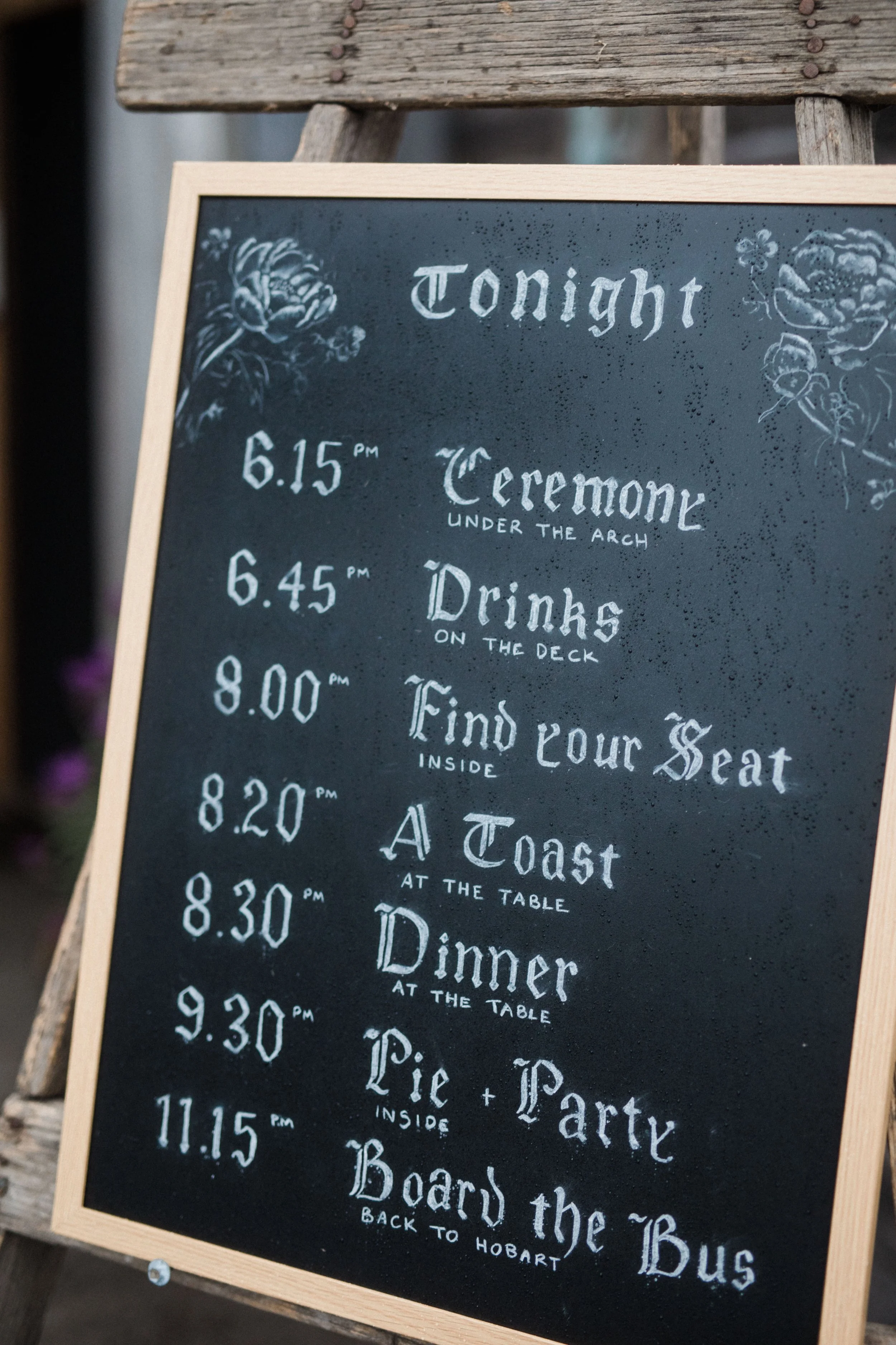 Event schedule on a chalkboard sign for tonight, including a ceremony at 6:15 PM, drinks on the deck at 6:45 PM, finding seats inside at 8:00 PM, a toast at the table at 8:20 PM, dinner at 8:30 PM, pie and party inside at 9:30 PM, and boarding the bu