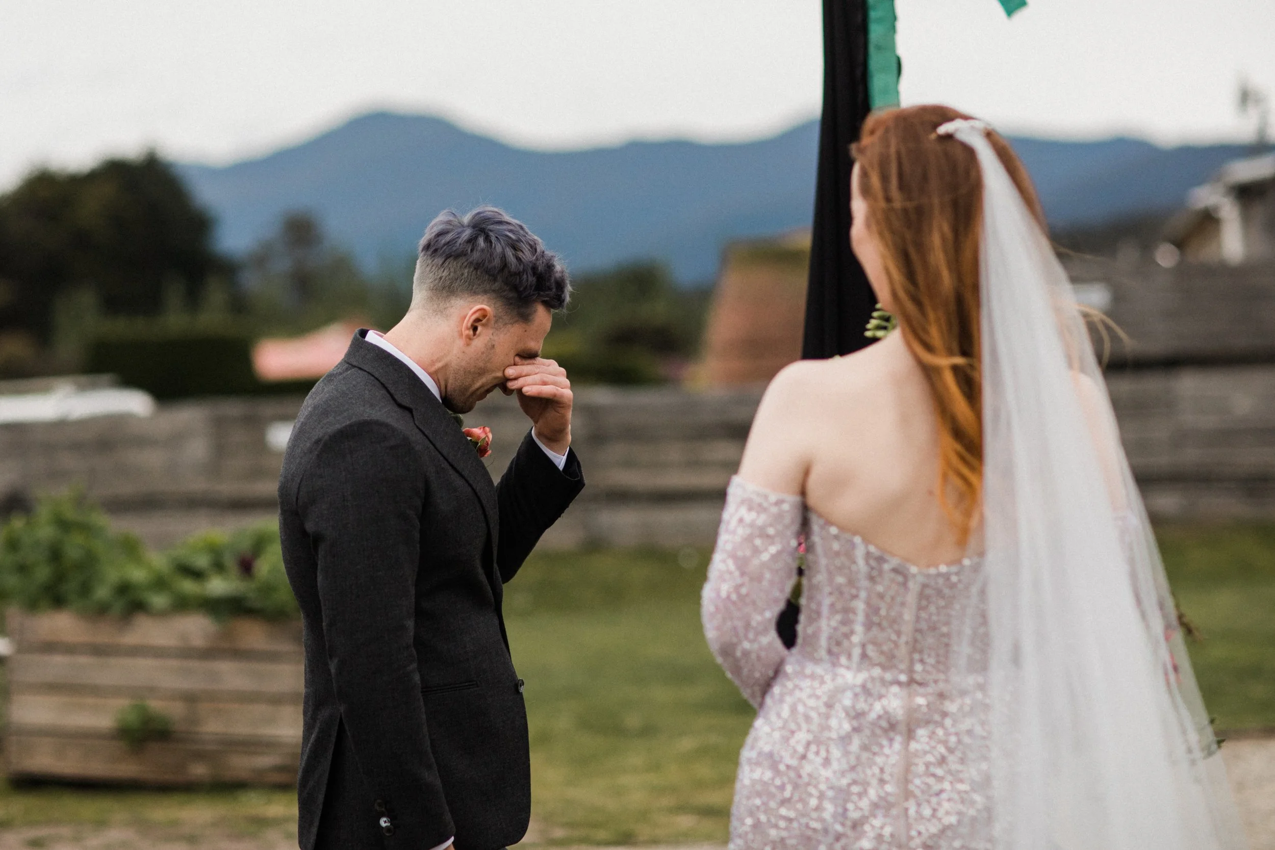A groom in a dark suit is emotional, covering his face with his hand, during a wedding ceremony outdoors. A bride with long red hair, wearing an off-shoulder lace wedding dress with a veil, faces him with her back to the camera. The background shows 