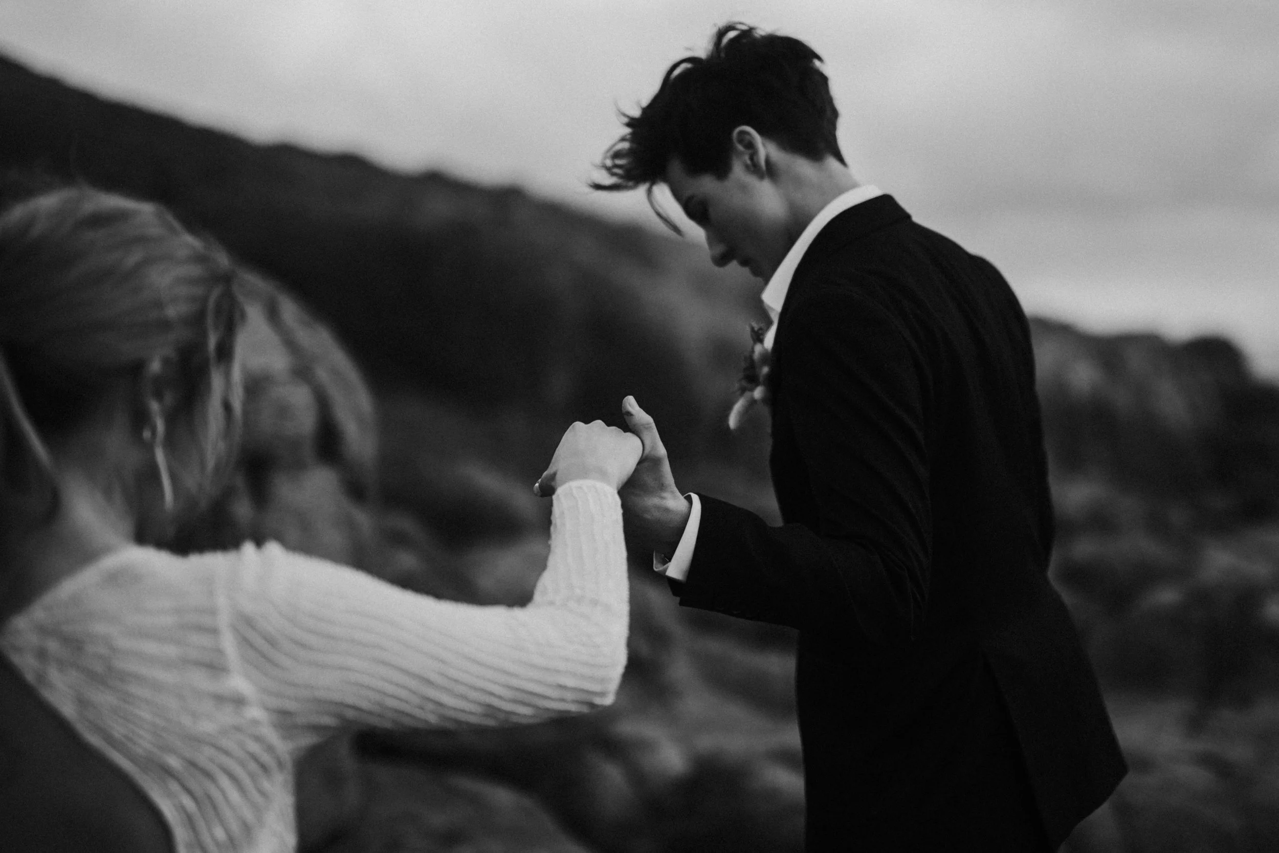 Black and white photo of a couple holding hands and gazing at each other outdoors with a mountainous landscape in the background.