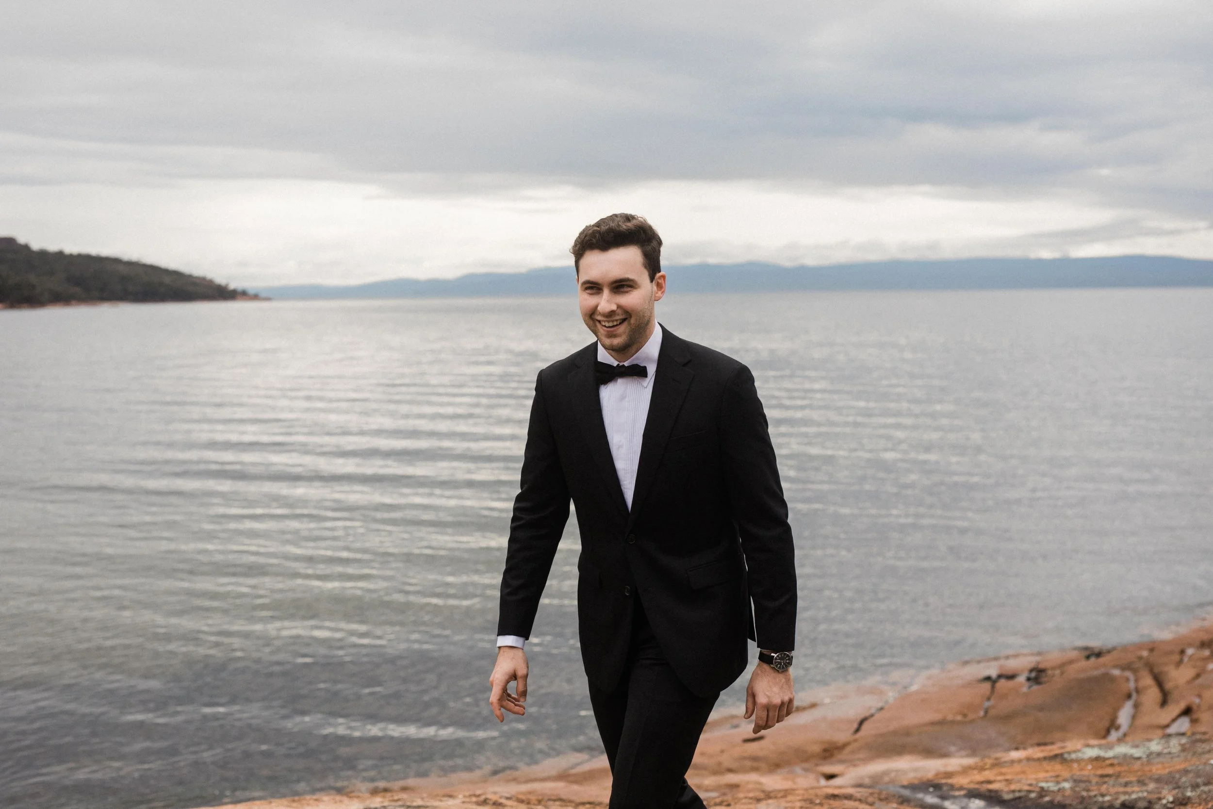 A young man in a black tuxedo with a bow tie walking on a rocky shoreline by a large body of water, with mountains in the background and a cloudy sky.
