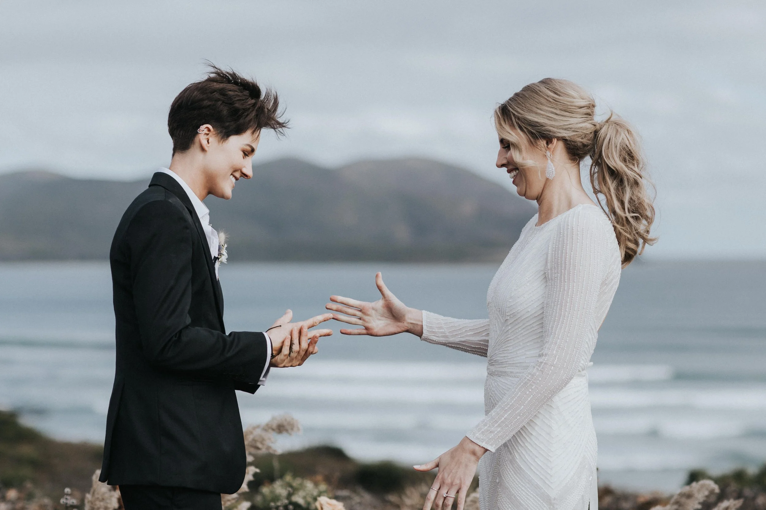 A couple during their wedding ceremony outdoors near the ocean, with mountains in the background, exchanging rings.