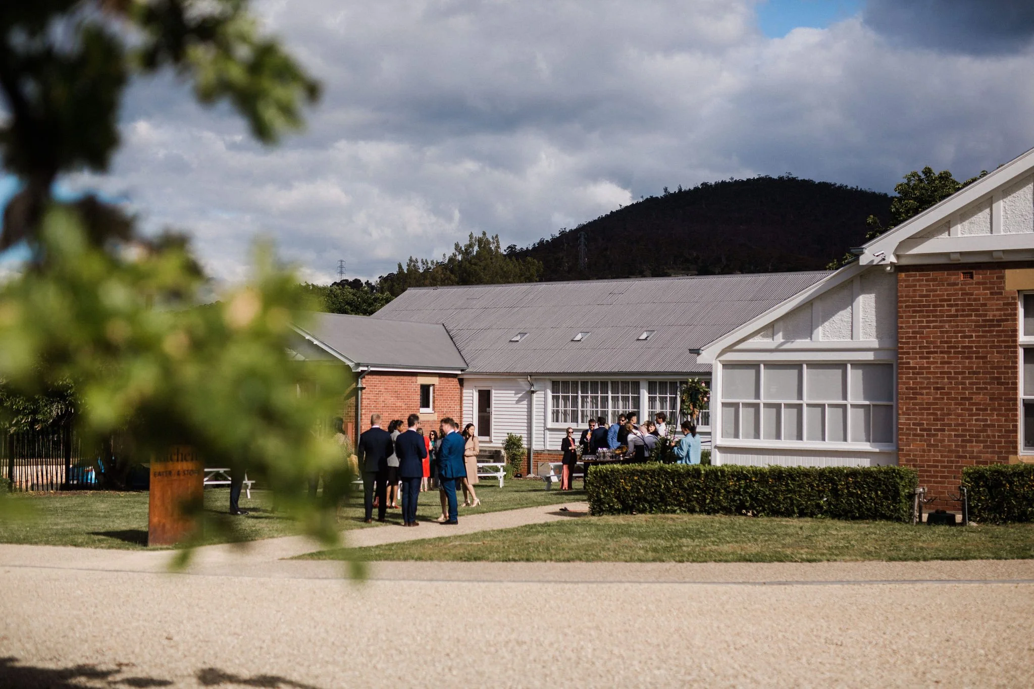 Group of people dressed formally gathered outside a building with gray roof and white walls, in a garden setting with mountains in the background.