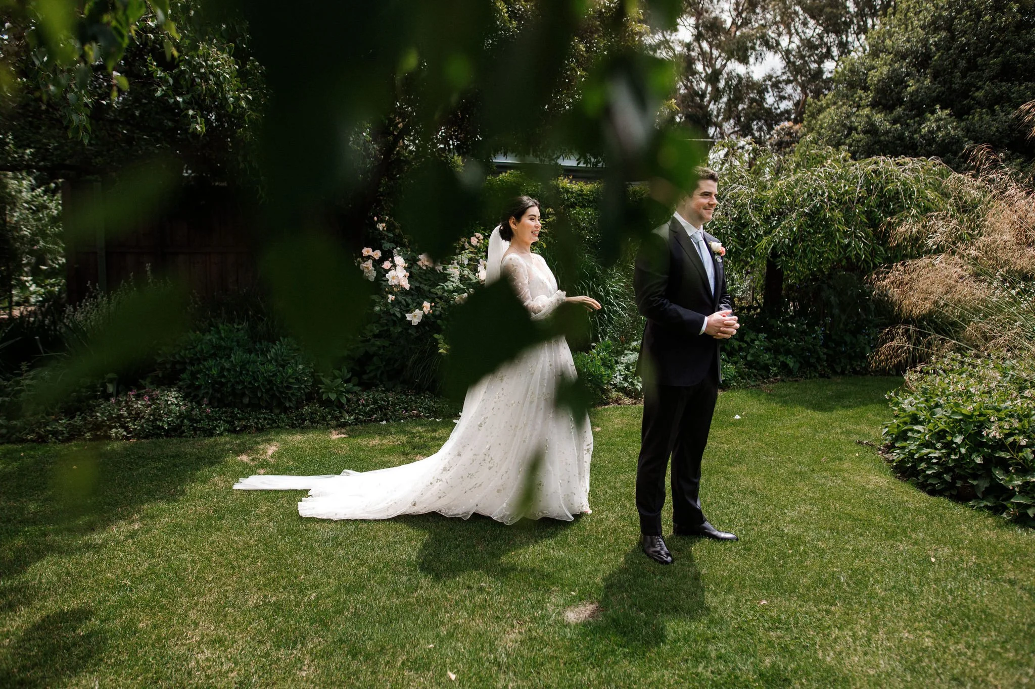 A bride and groom stand outdoors on a lush green lawn surrounded by trees and flowering bushes, with the bride smiling behind some leaves.