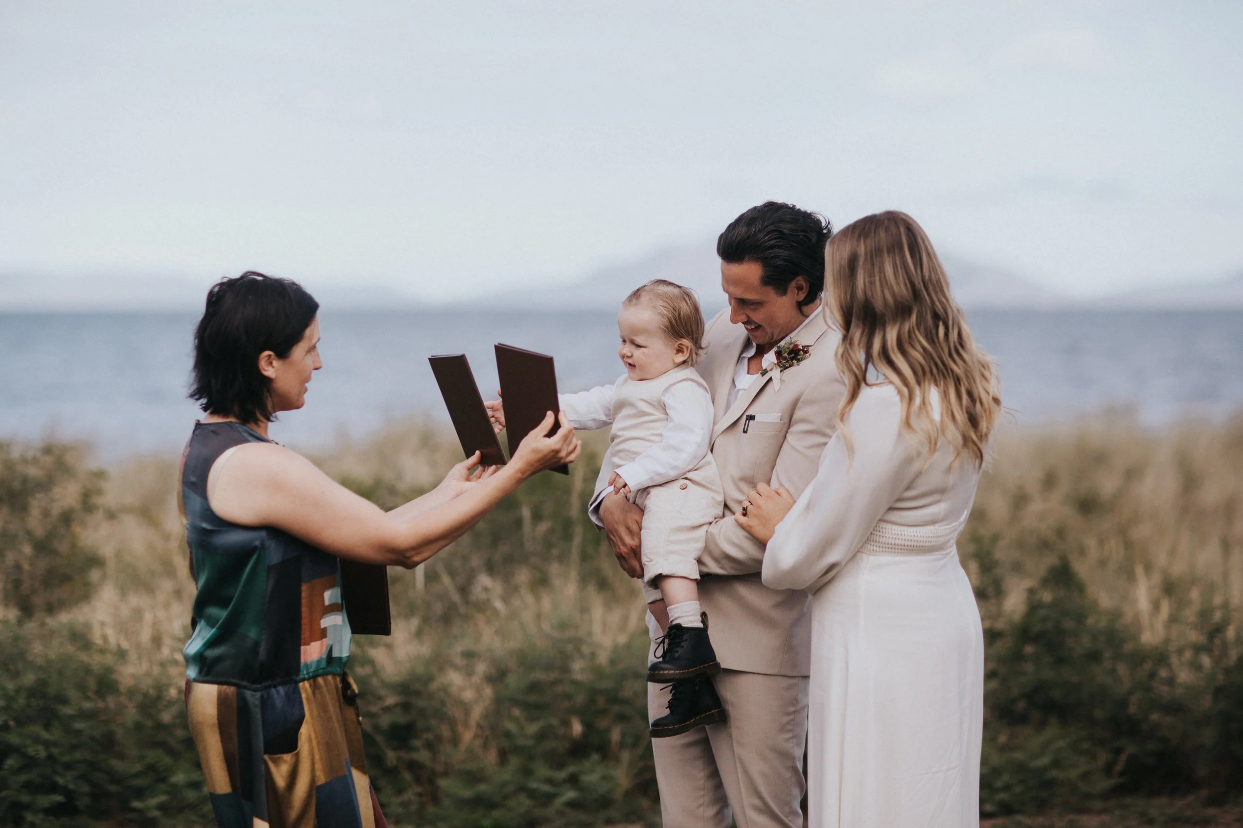 A couple at a beach wedding exchanging rings with a woman officiating, while holding a child between them.
