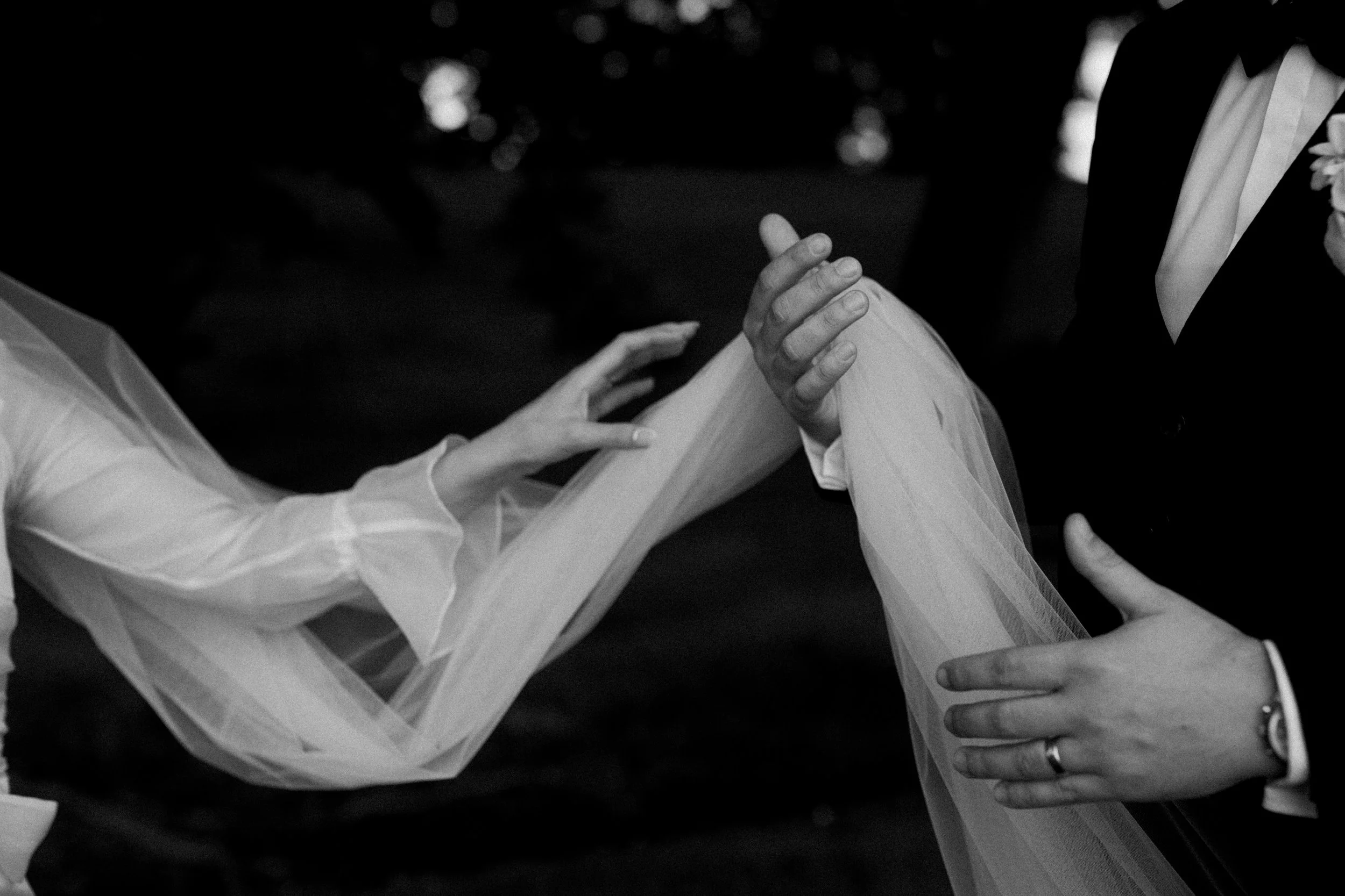 Close-up black and white photo of two people at a wedding, with one person wearing a light-colored dress and the other in a traditional suit with a boutonniere, holding hands with a veil softly flowing between them.