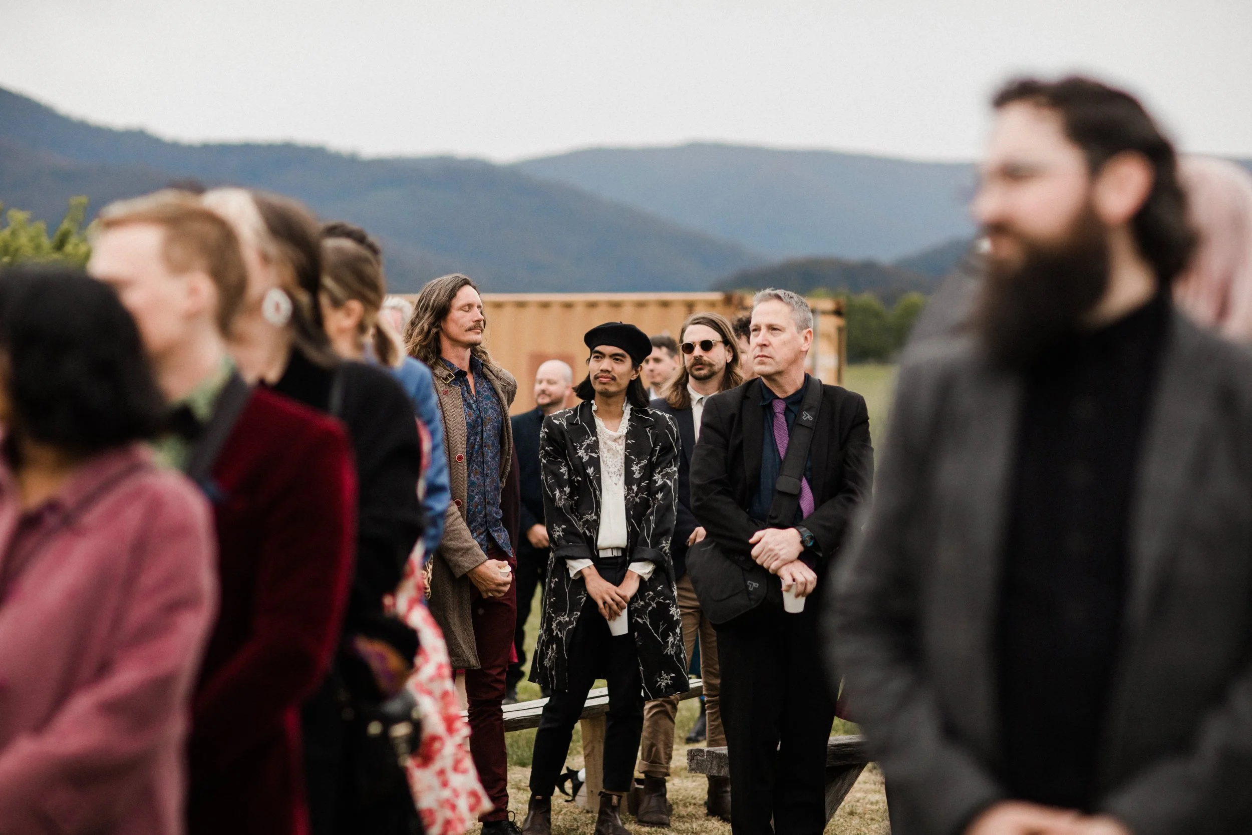 A group of people standing outdoors in a line, some engaged with their surroundings, with mountains in the background.