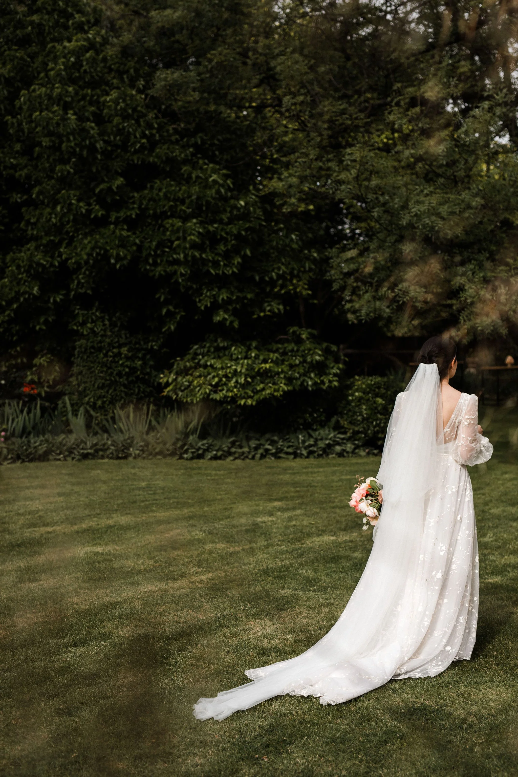 A bride in a white wedding dress with a long train and veil, holding a bouquet of flowers, standing on a grassy lawn with trees in the background, facing away from the camera.