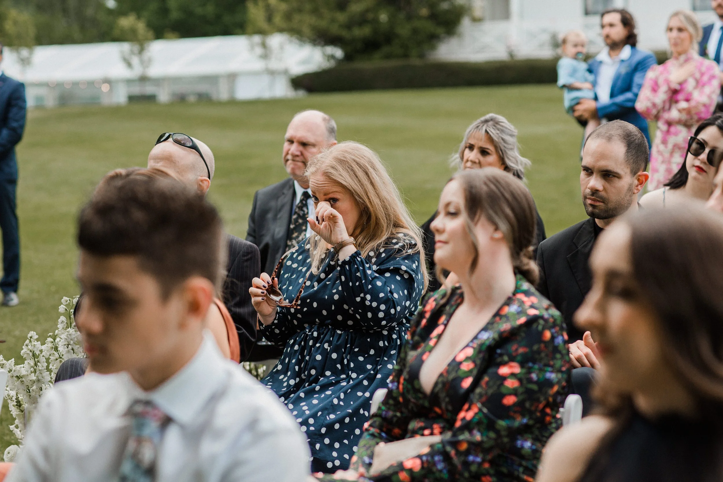 People seated outdoors at a formal event, with a woman in a blue polka dot dress wiping tears from her eyes.