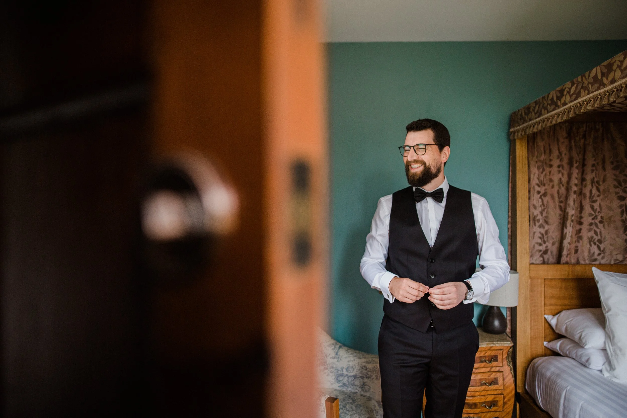 A groom preparing for his wedding, adjusting his suit, standing in a bedroom with a wooden bed and teal wall, smiling.