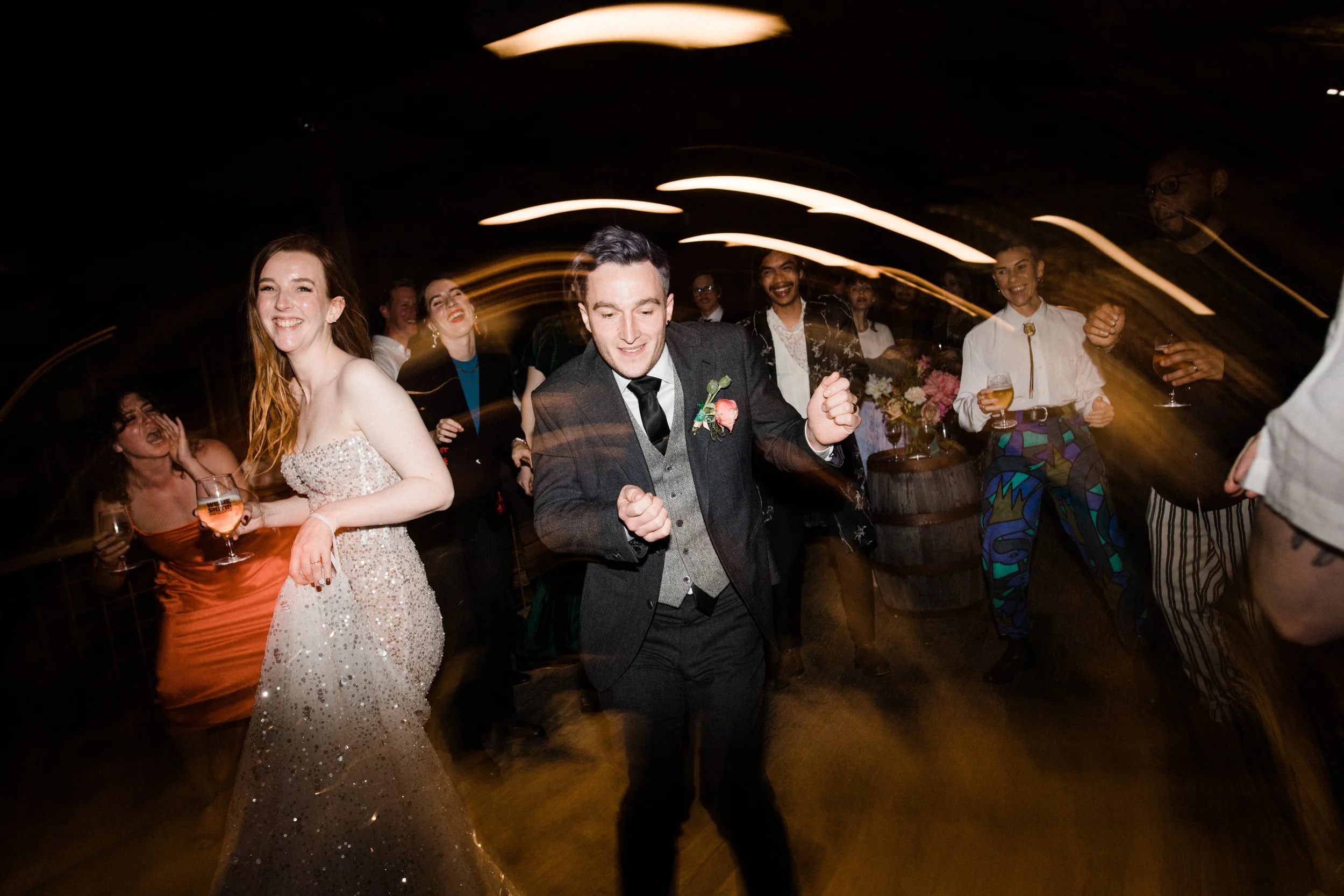 People dancing and celebrating at a wedding reception, with a man in a suit and a woman in a sparkling dress in the foreground, surrounded by guests holding drinks and smiling.