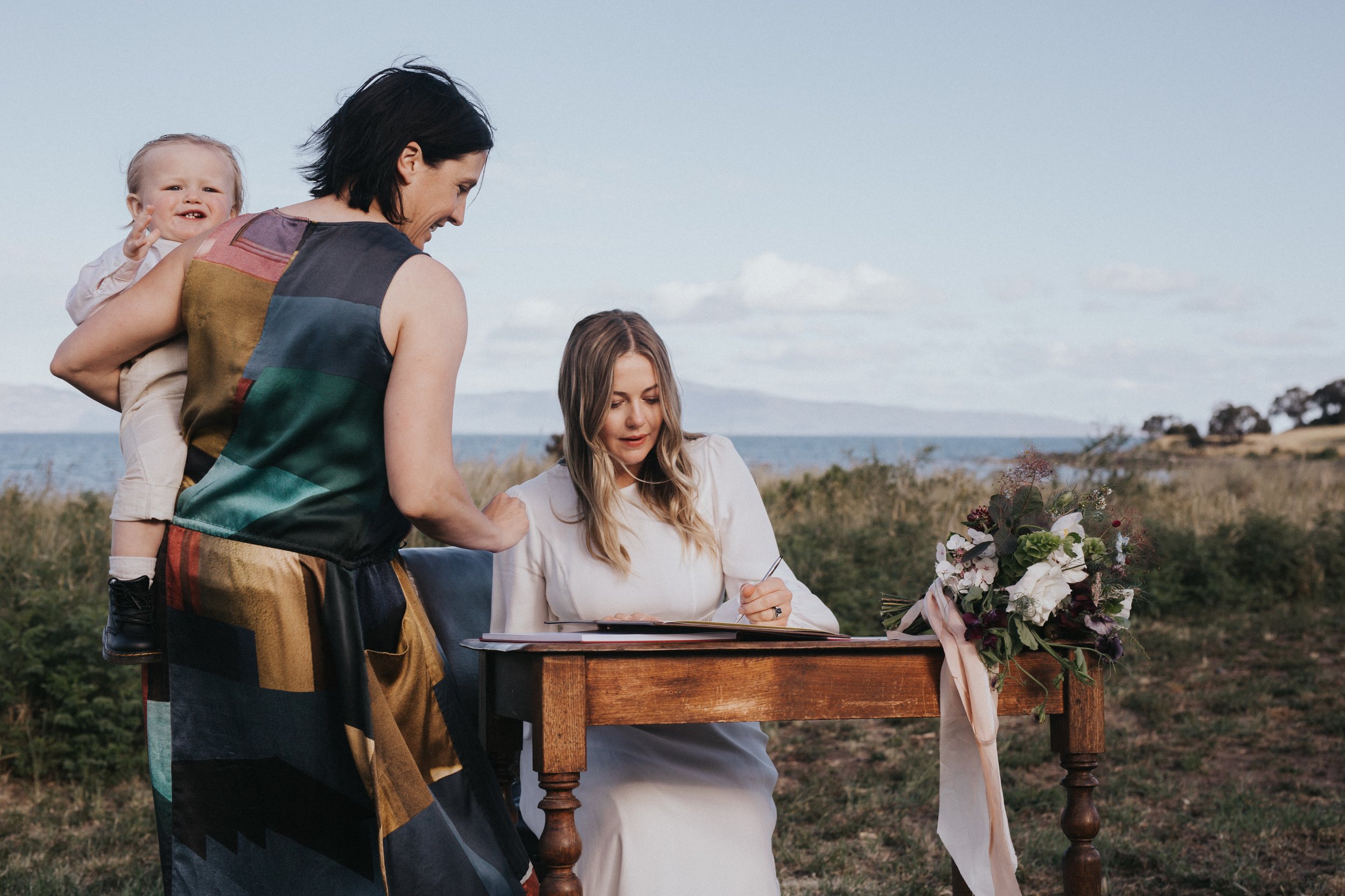 A woman is signing a book at a wedding ceremony outdoors near the sea, with a flower bouquet on the table, a woman with a child standing behind her.