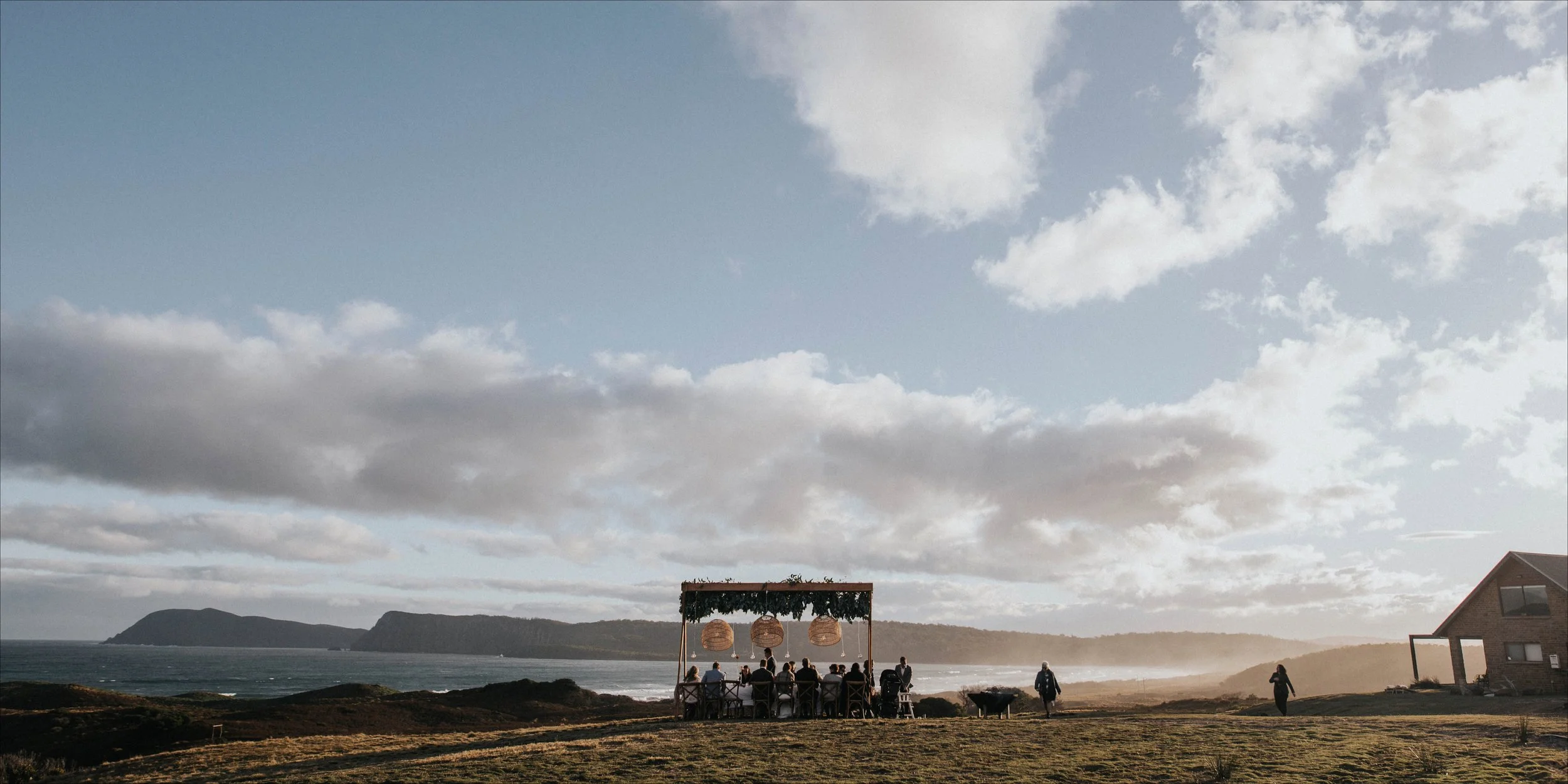 Guests seated at a dining table outdoors near the coast with ocean and hills in the background, under a partly cloudy sky.