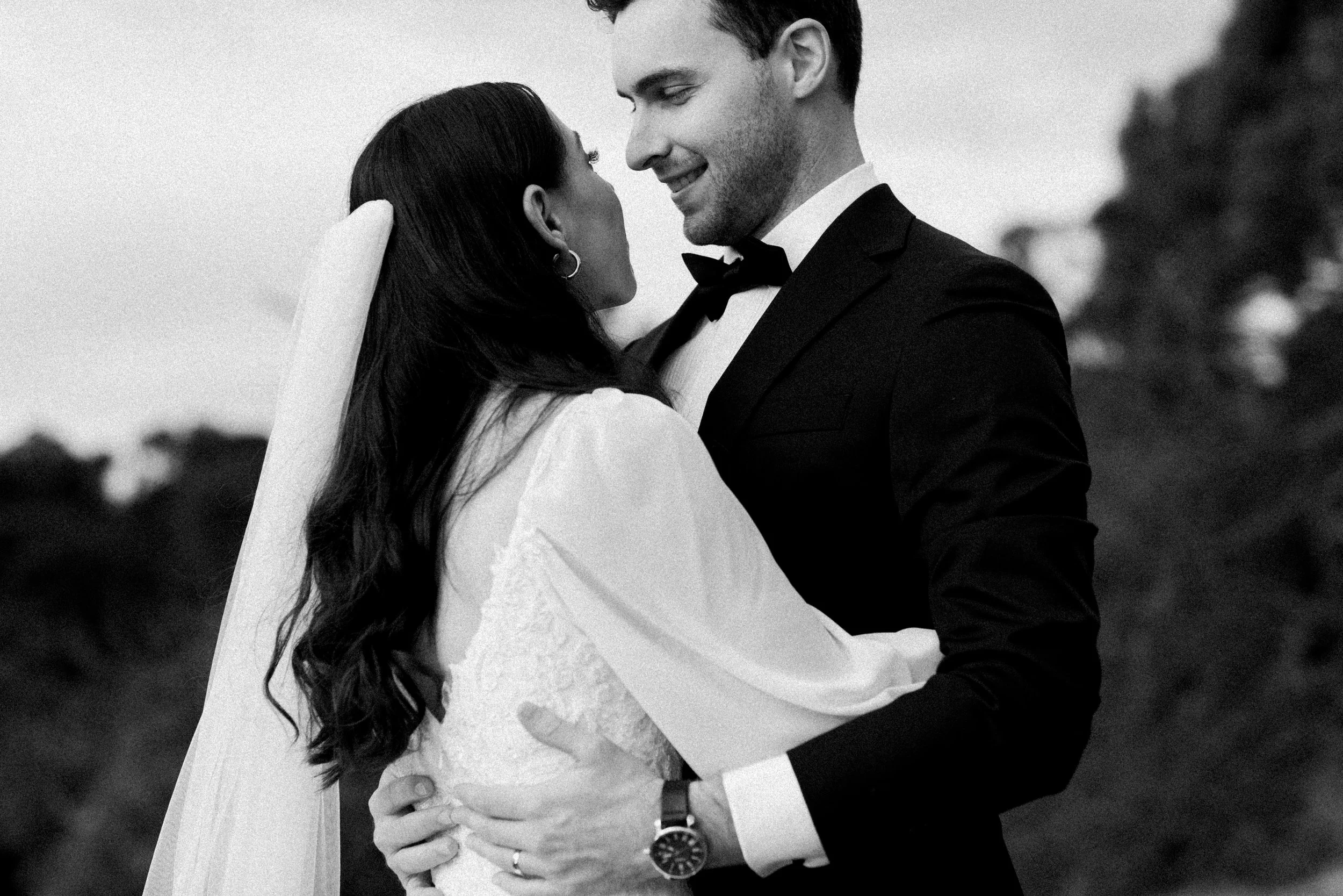 A black and white photo of a bride and groom embracing, smiling at each other, with the bride wearing a veil and lace dress, and the groom in a tuxedo, outdoors with blurred trees in the background.