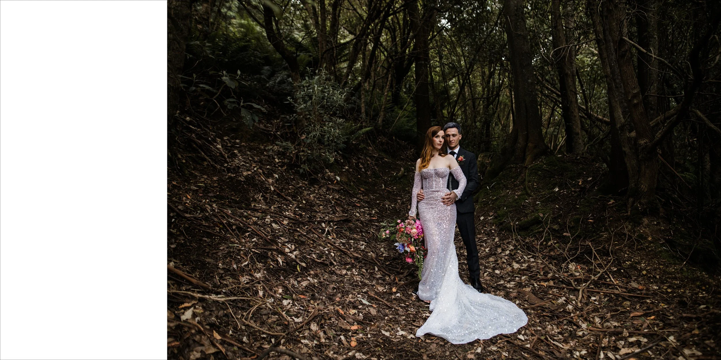 A bride and groom standing in a wooded area for a wedding photo. The bride is in a long, sparkly, off-the-shoulder wedding dress holding a bouquet of pink and purple flowers. The groom is in a black tuxedo with a boutonniere. They are close together,