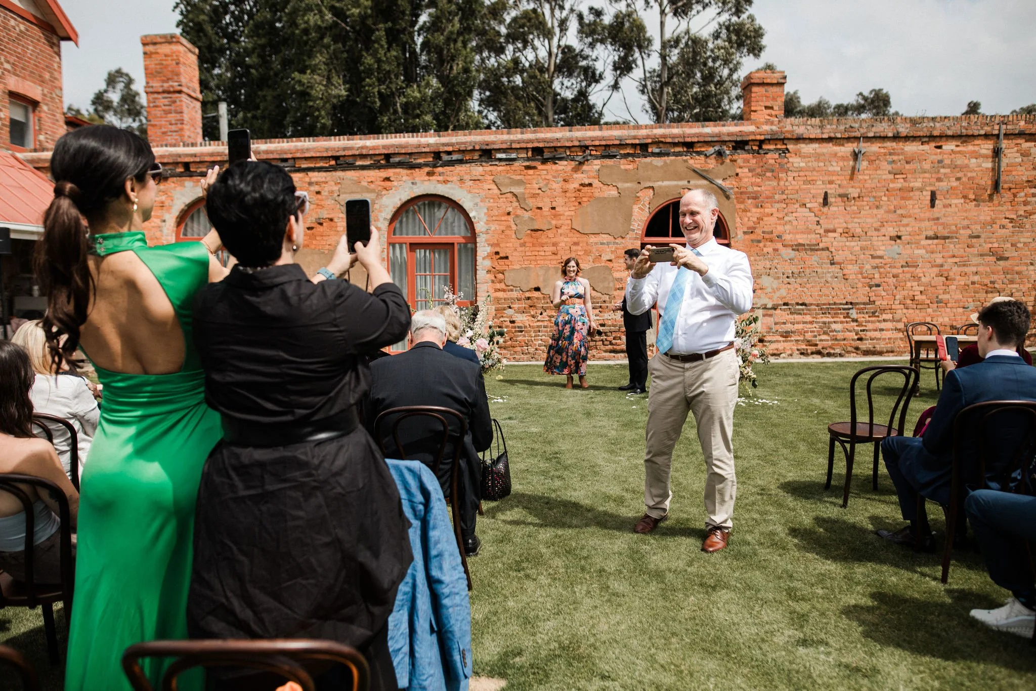 Man in white shirt and beige pants taking a photo of a woman in floral dress at outdoor wedding ceremony on a grassy area in front of a brick wall.