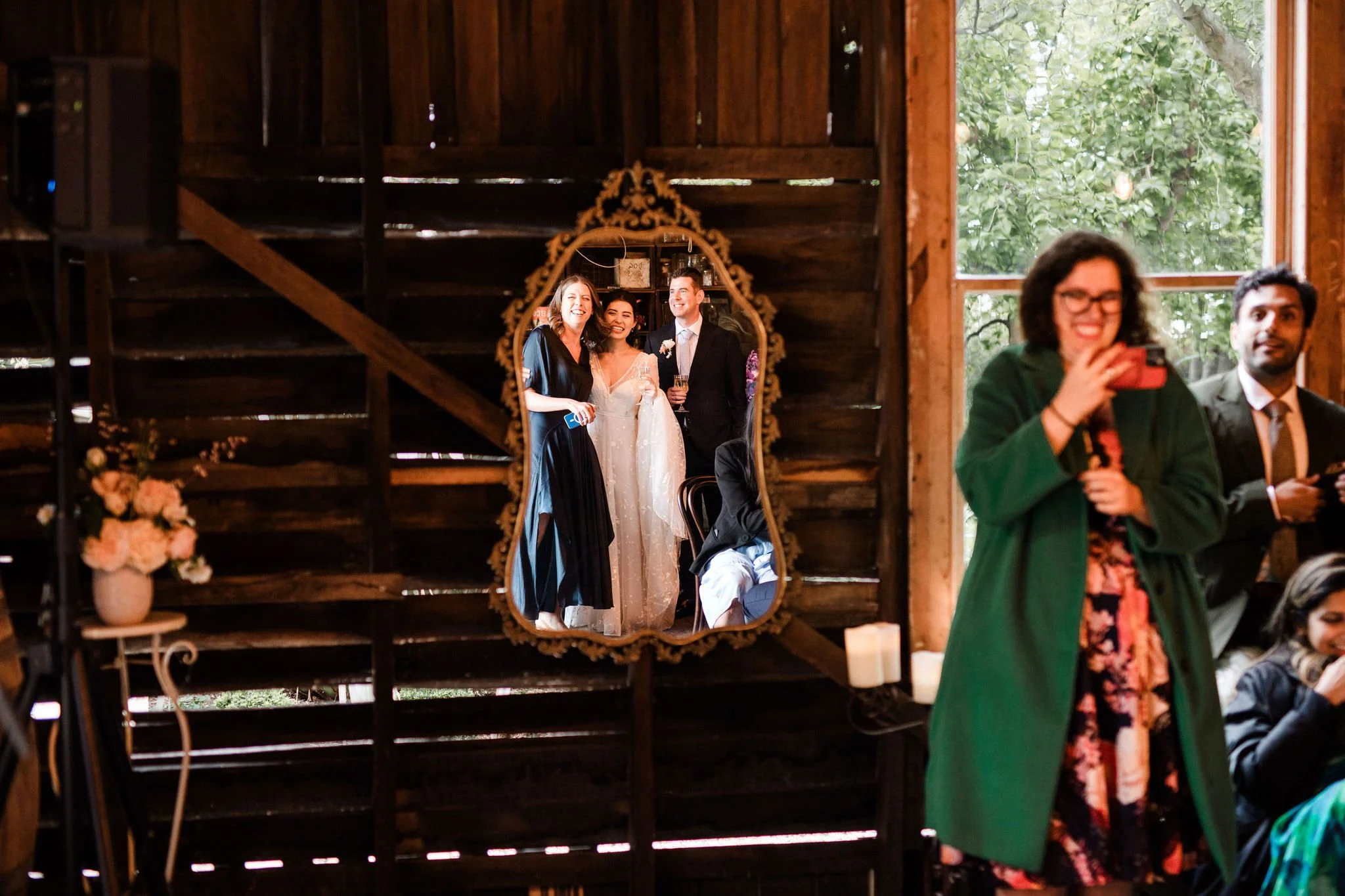 A mirror reflecting a bride and groom with two women celebrating at a rustic wedding reception, with guests and natural light from a window nearby.