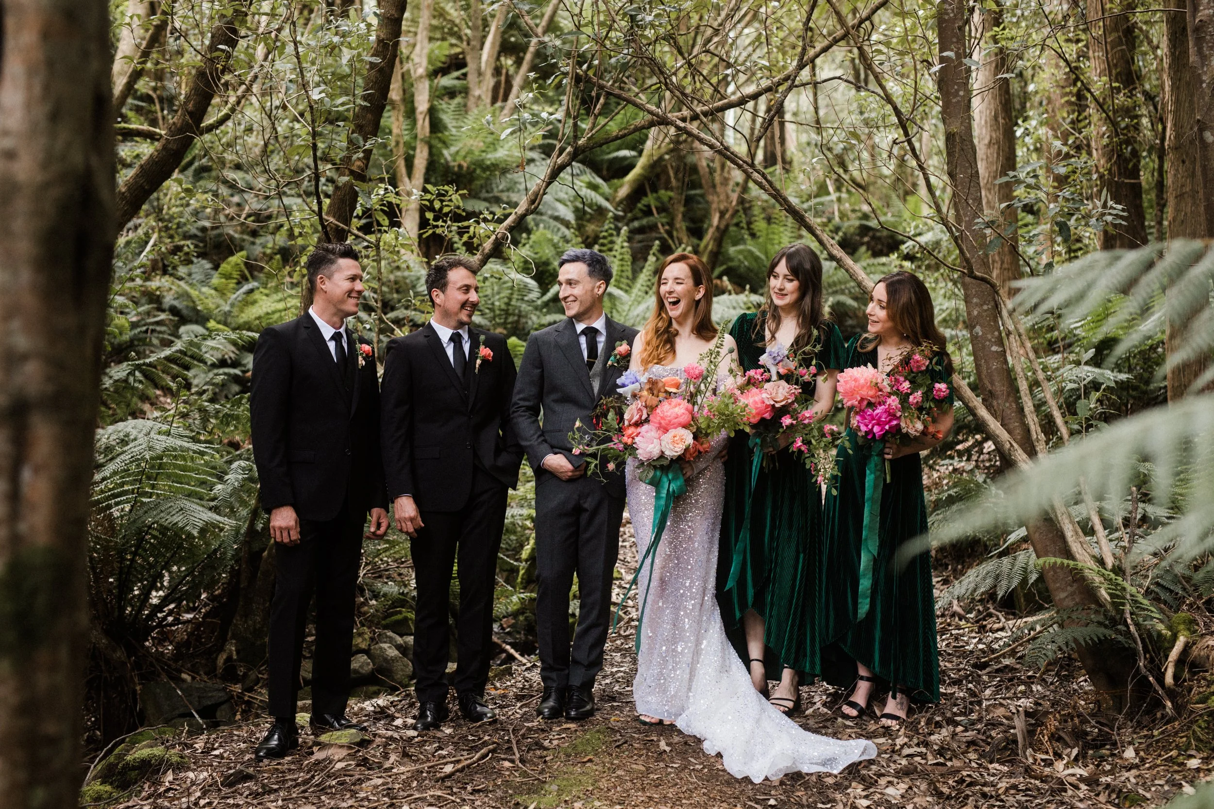 A wedding party standing in a lush forest, with the bride holding a bouquet of pink and purple flowers and the bridesmaids holding similar bouquets. The men are dressed in black suits and ties. Everyone appears joyful and is smiling.