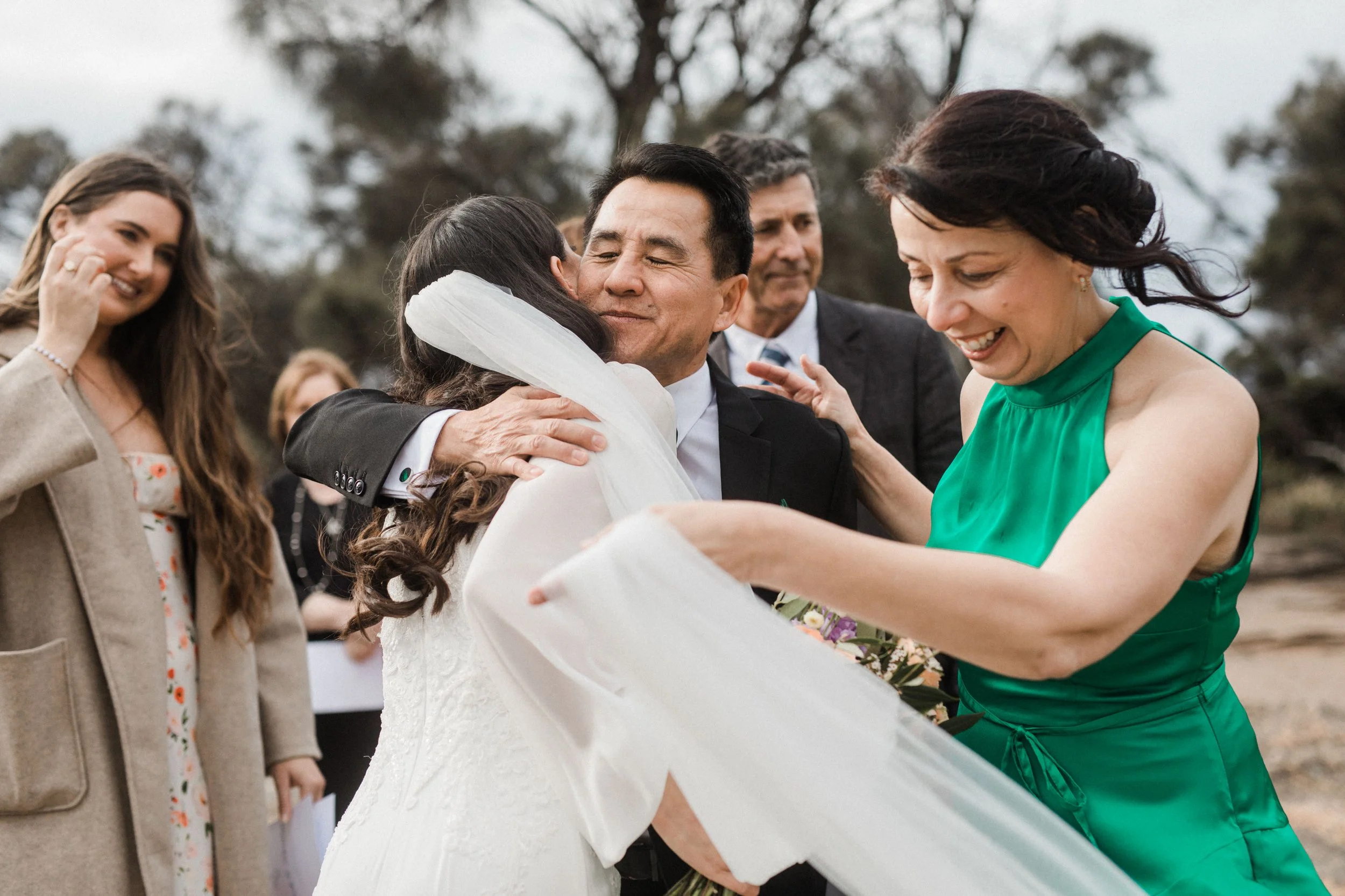 People embracing and celebrating at a wedding ceremony outdoors, with a woman in a green dress and a bride in a white gown hugging.