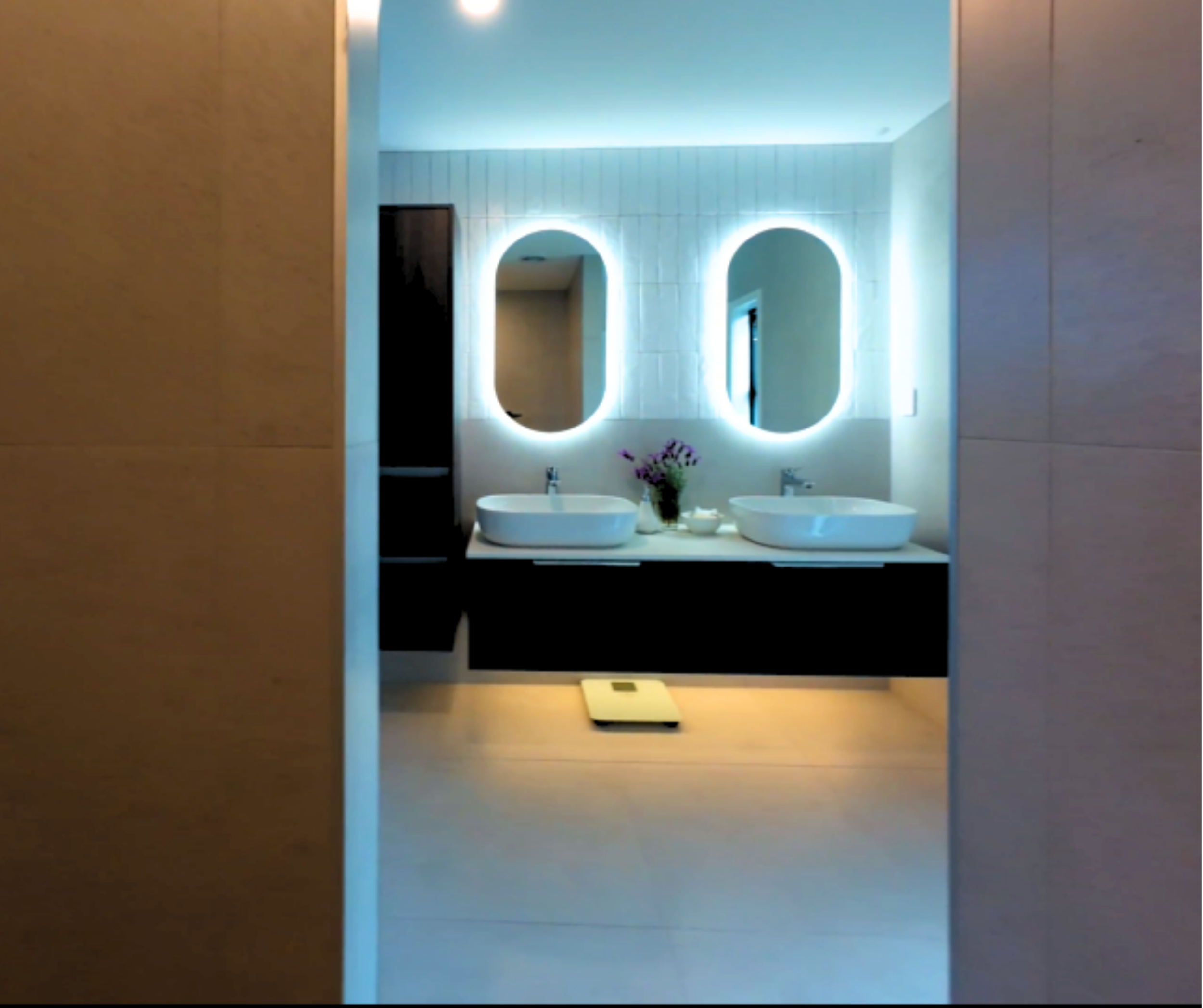 Modern bathroom with two vessel sinks, illuminated oval mirrors, a black vanity, a scale on the floor, and a vase of purple flowers between the sinks. Volt Solutions electrician installed the electrics in this room. 