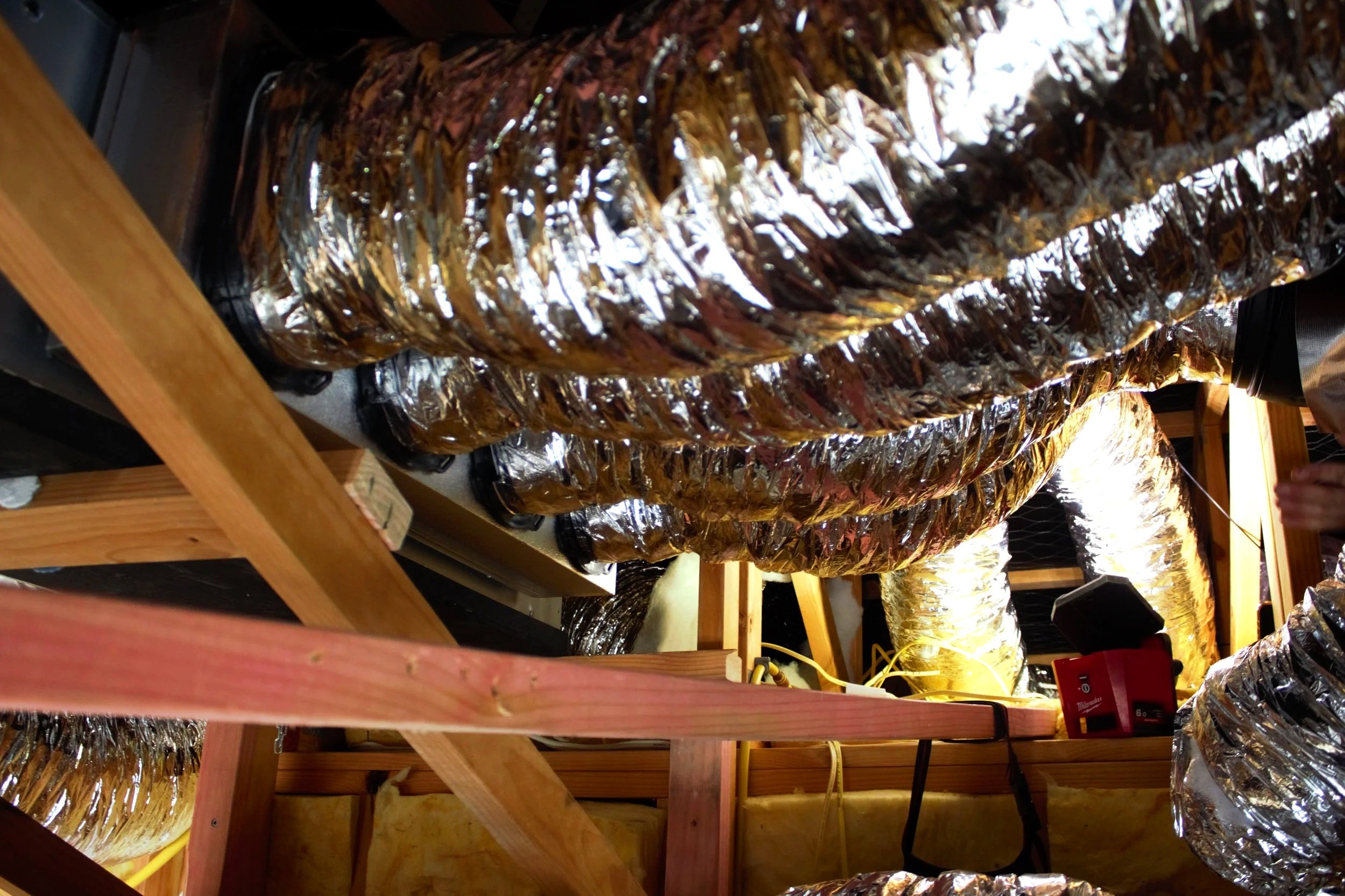 Multiple shiny, metallic ducts suspended in an attic space with wooden framing, yellow insulation, and electrical cords. Volt Solutions electrician installed this.