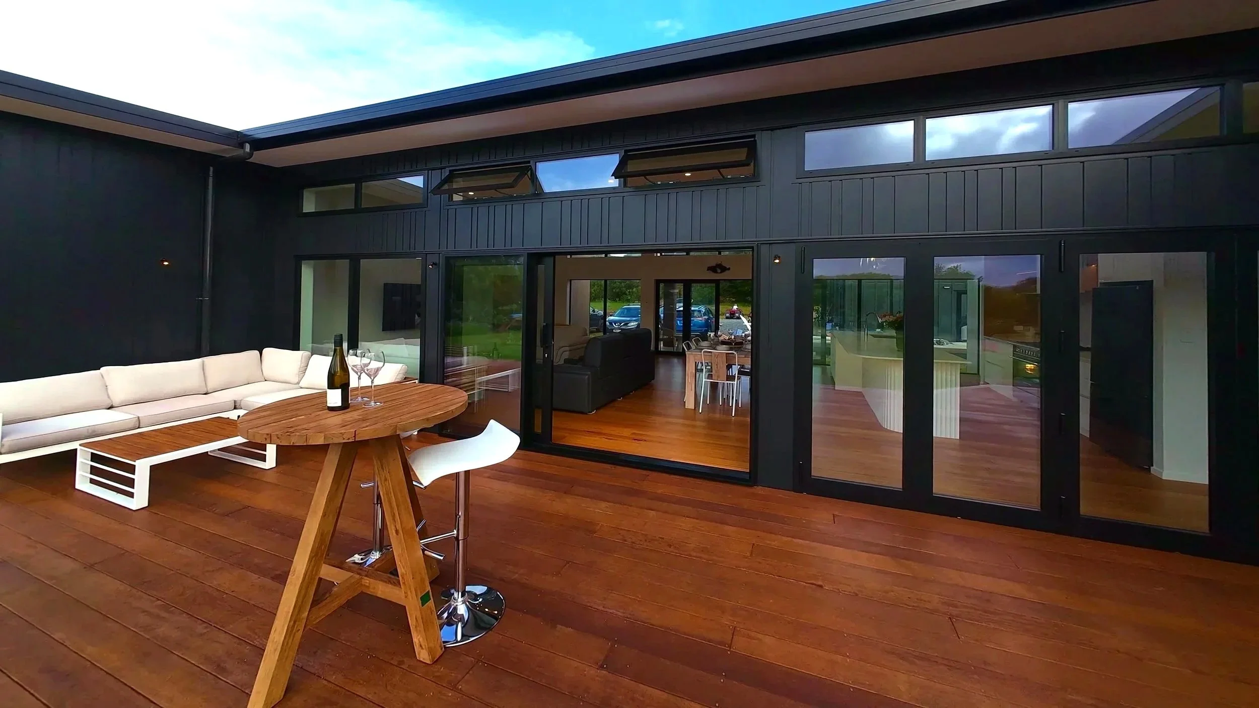 Modern house with a large outdoor wooden deck, black exterior walls, sliding glass doors, white and black furniture, a wine bottle, and open windows. Electrical installed by Volt Solutions electrician.