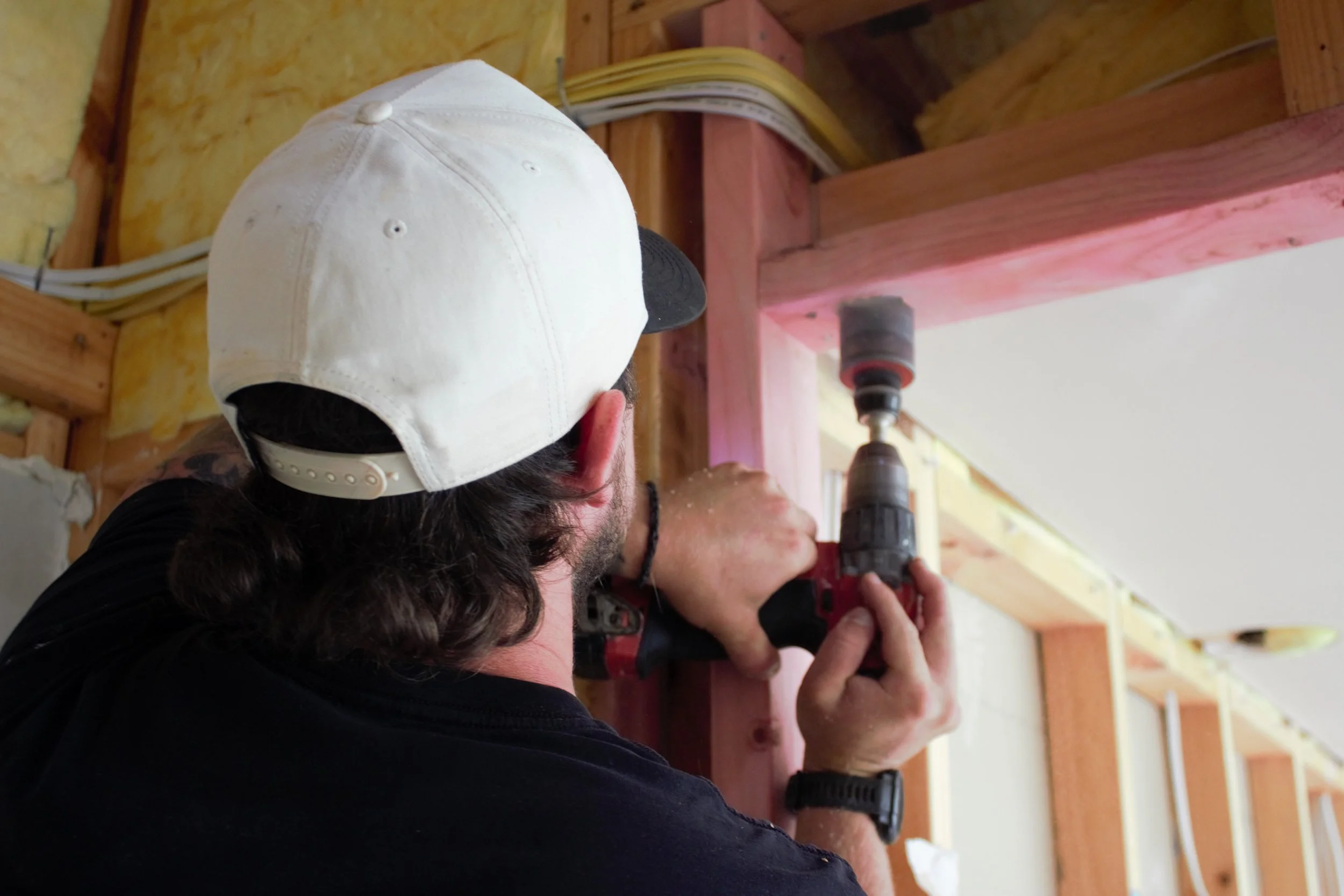  Volt Solutions electrician with long dark hair wearing a white cap and black shirt is using a power drill to drill into wooden beams in an indoor construction or renovation setting.