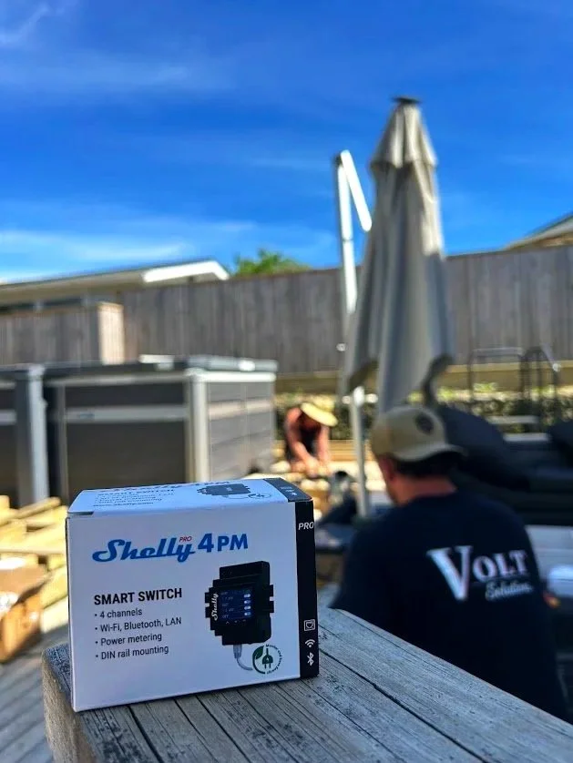 A boxed Shelly 4PM smart switch placed on a wooden surface outdoors in a backyard. Dan in a black shirt and a beige hat is working with other equipment, and another person is kneeling near a garden bed with a large umbrella, fencing, and a blue sky.